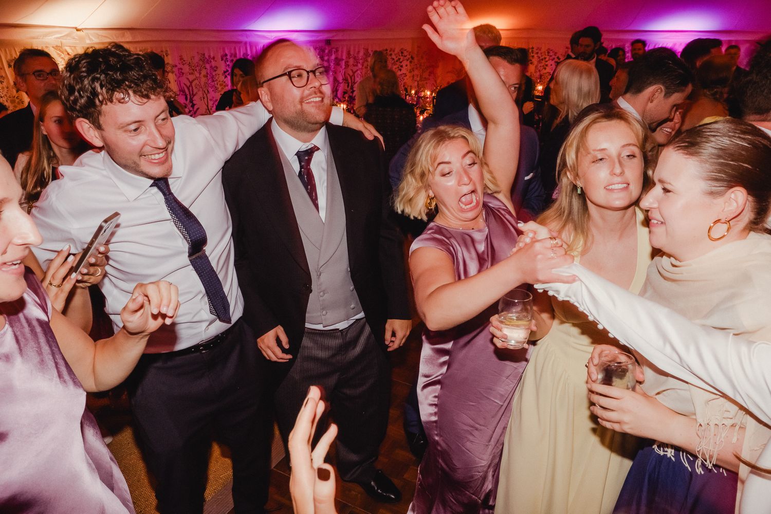 Wedding guests dance and celebrate on a colorful dance floor illuminated by purple and orange lighting at a reception.