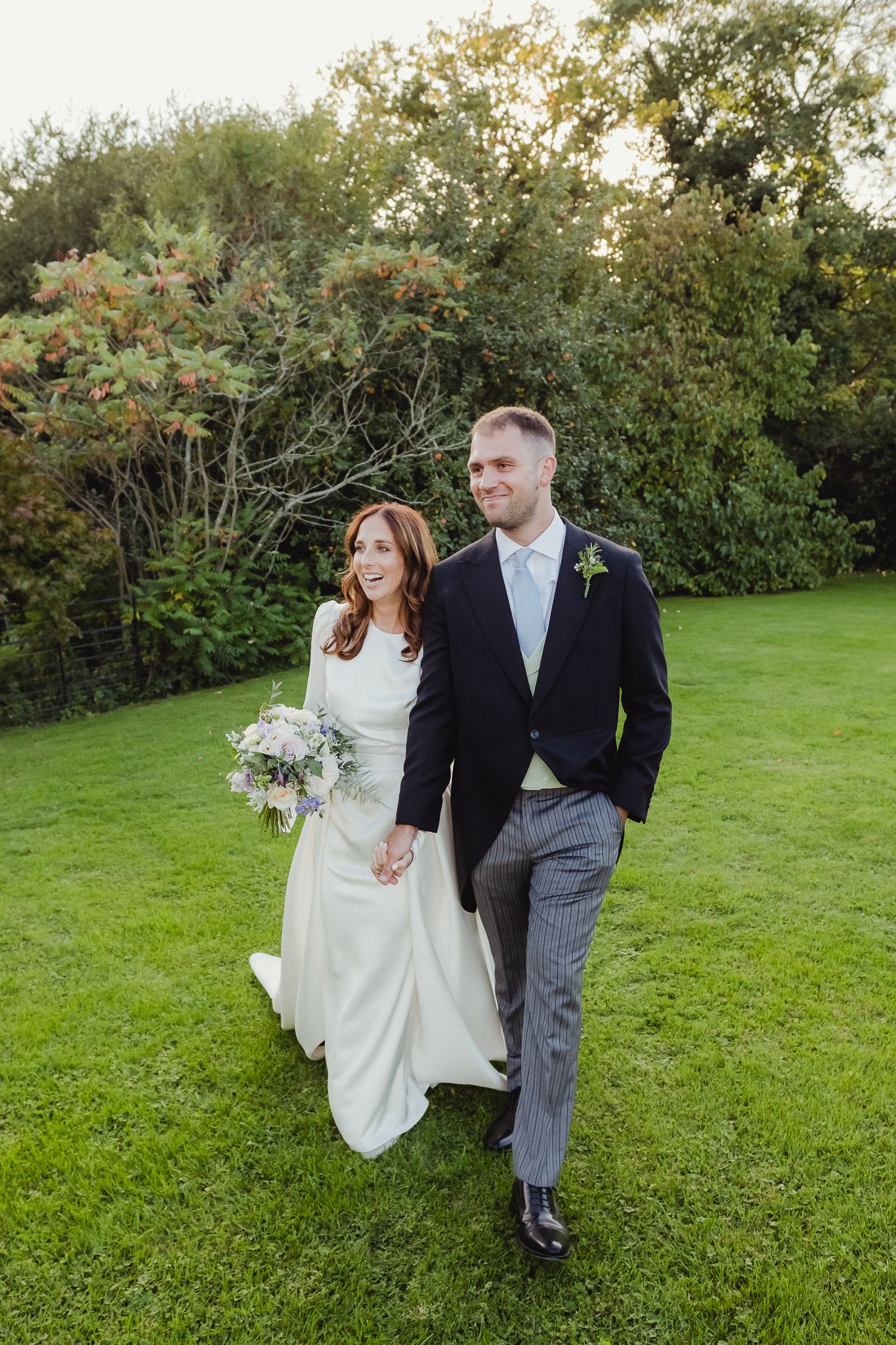 A couple in formal wedding attire walks together across a lush green lawn on their special day.