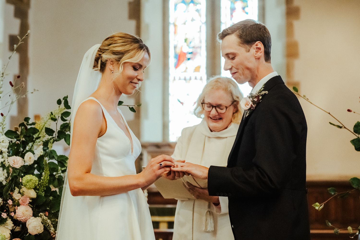 Wedding ceremony in sunlit church with floral arrangements and stained glass windows.