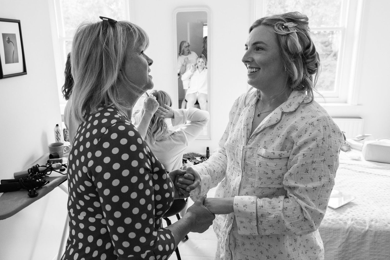 Two women hold hands and smile warmly in a bright room, one in a polka dot blouse and the other in pajamas, in a black and white photo.