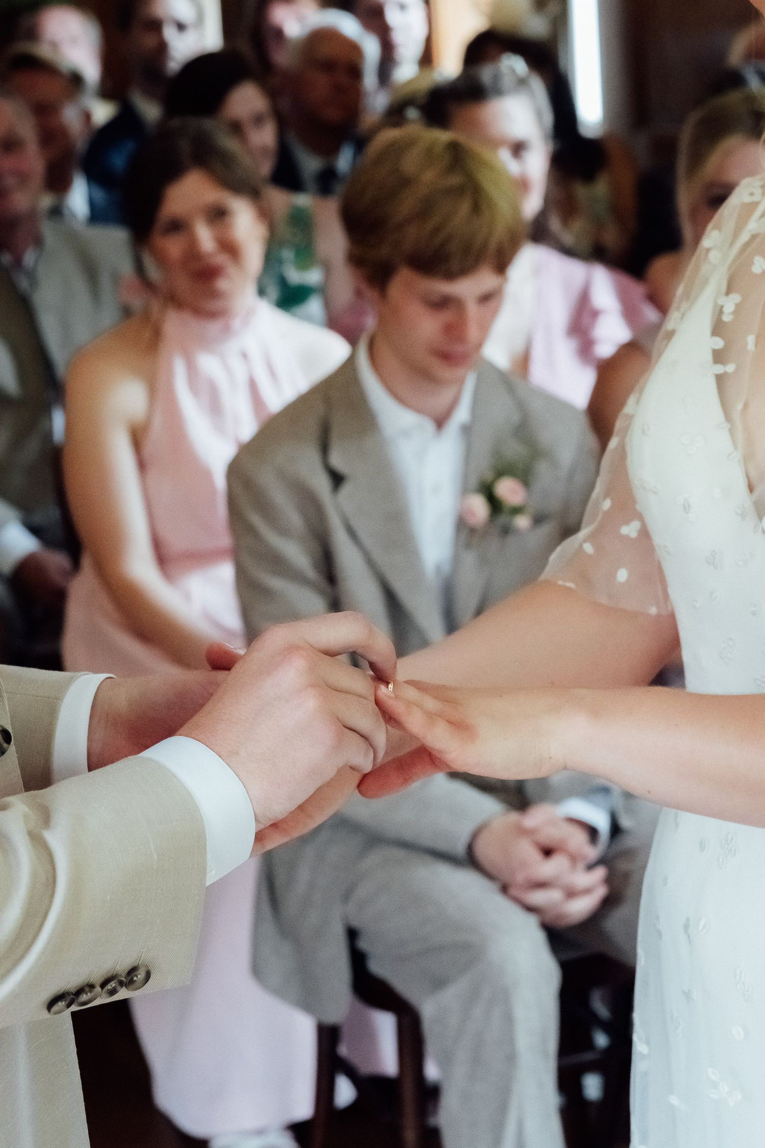 Bride in a white lace gown receives a wedding ring during the ceremony as smiling guests in pastel and gray attire look on.