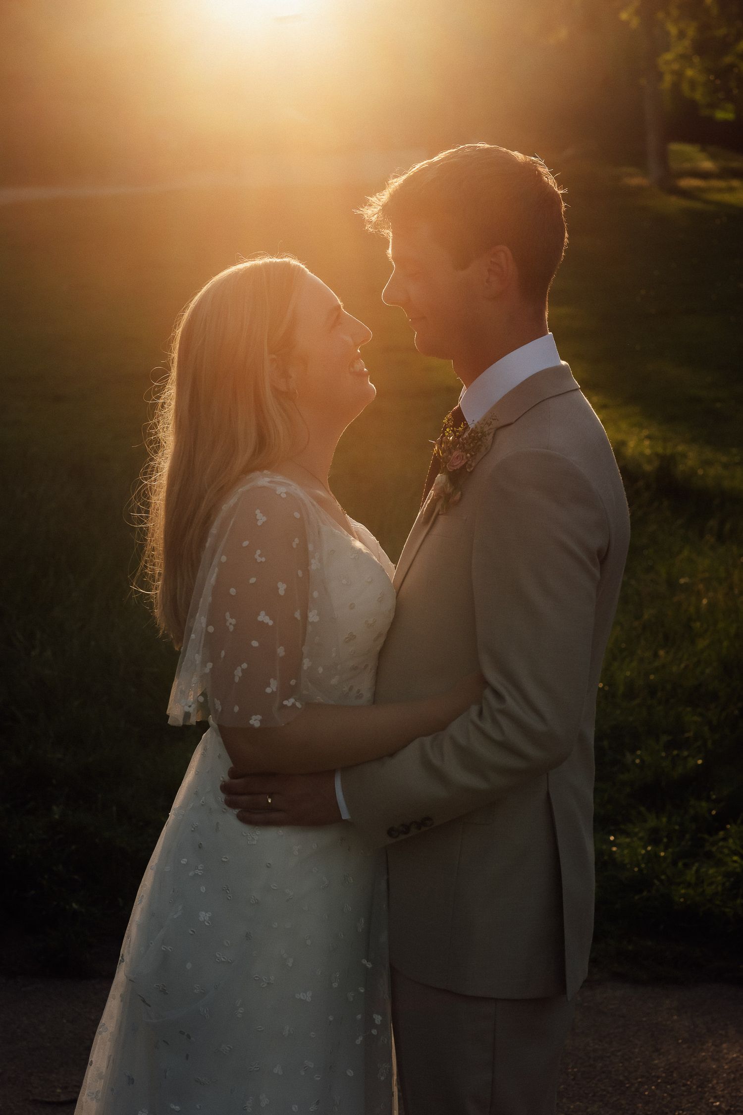 A bride in a white lace dress and a groom in a tan suit embrace face to face, glowing in golden sunset light at their outdoor wedding.