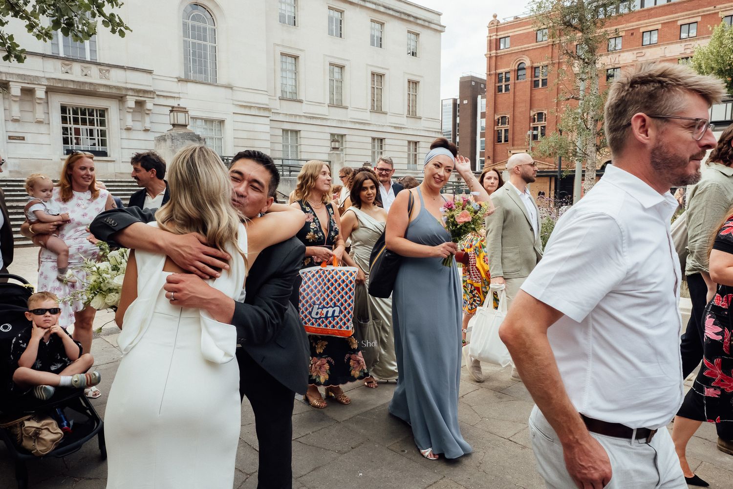 Bride in a white dress embraces a groom outside while wedding guests in formal attire gather and celebrate on a sunny city street.