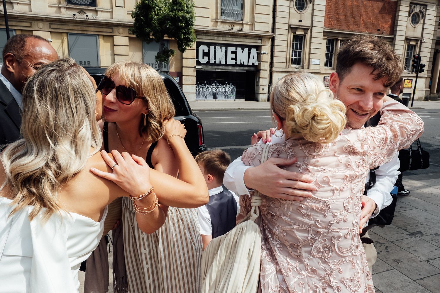 Wedding guests in summer outfits share warm hugs and smiles on a sunny street outside a building with a Cinema sign.