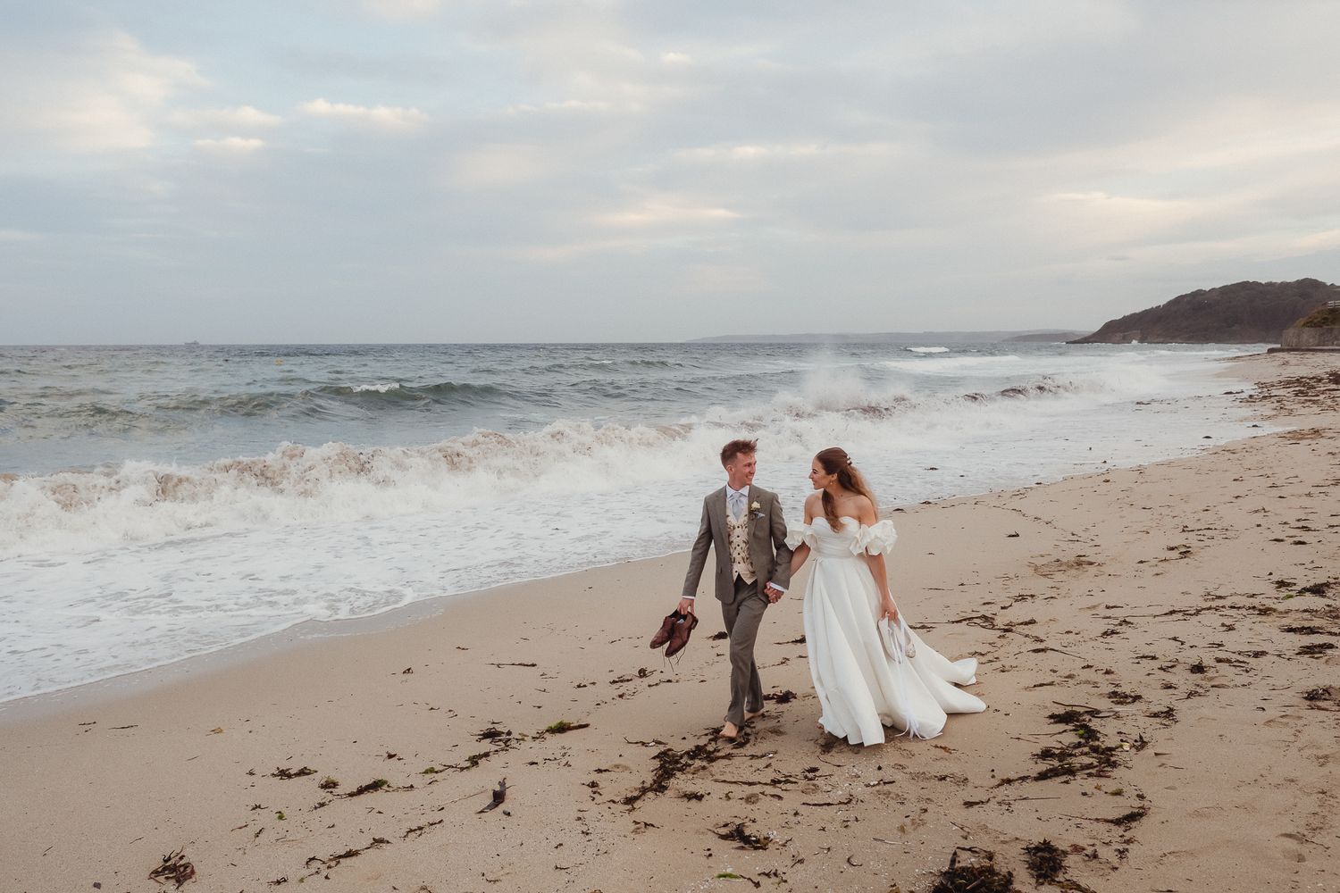 A couple walks along a scenic beach shoreline with waves crashing against the sand during a cloudy day.