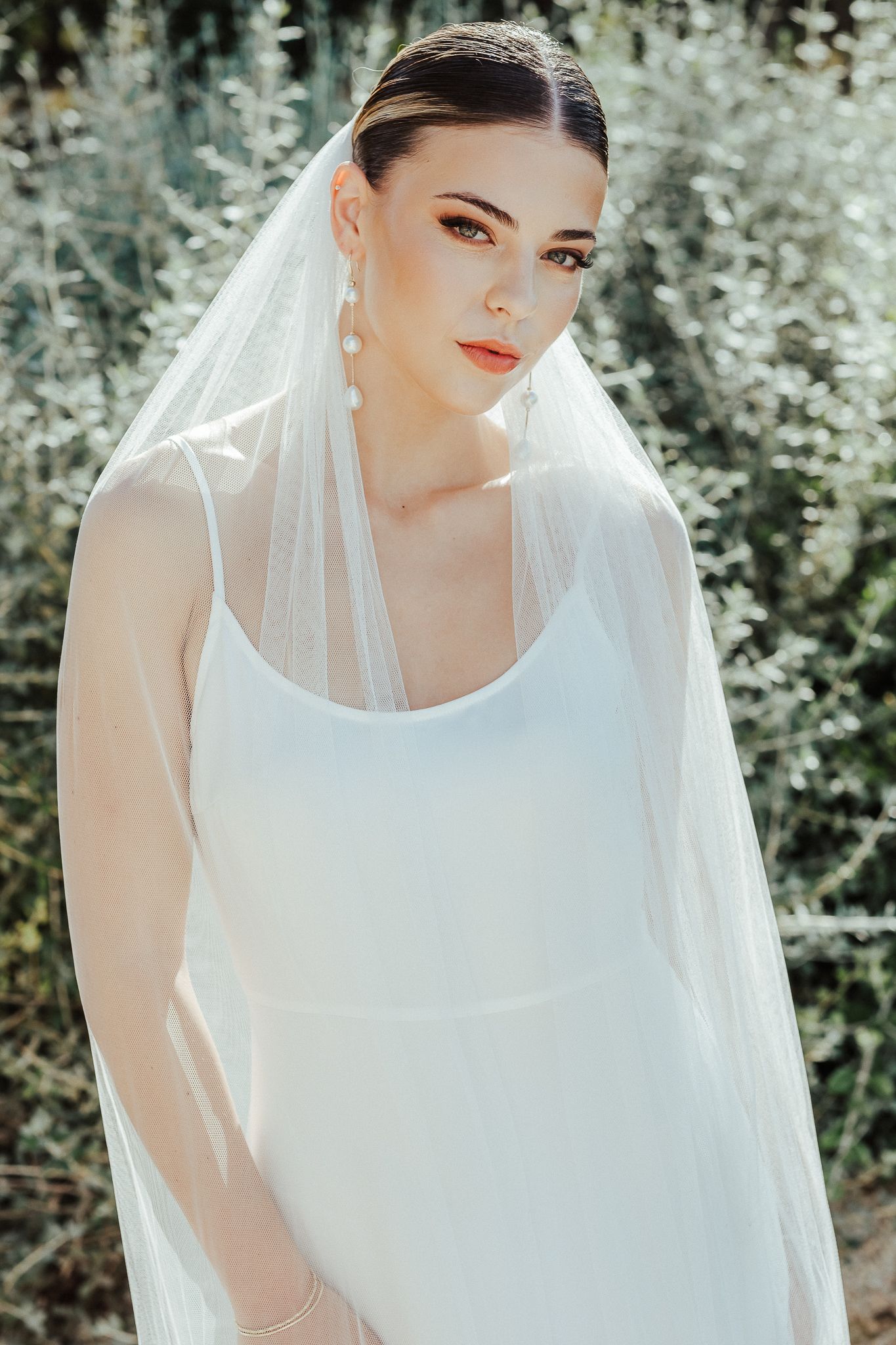 A bride in a white wedding dress and veil poses outdoors against a soft, blurred natural background.
