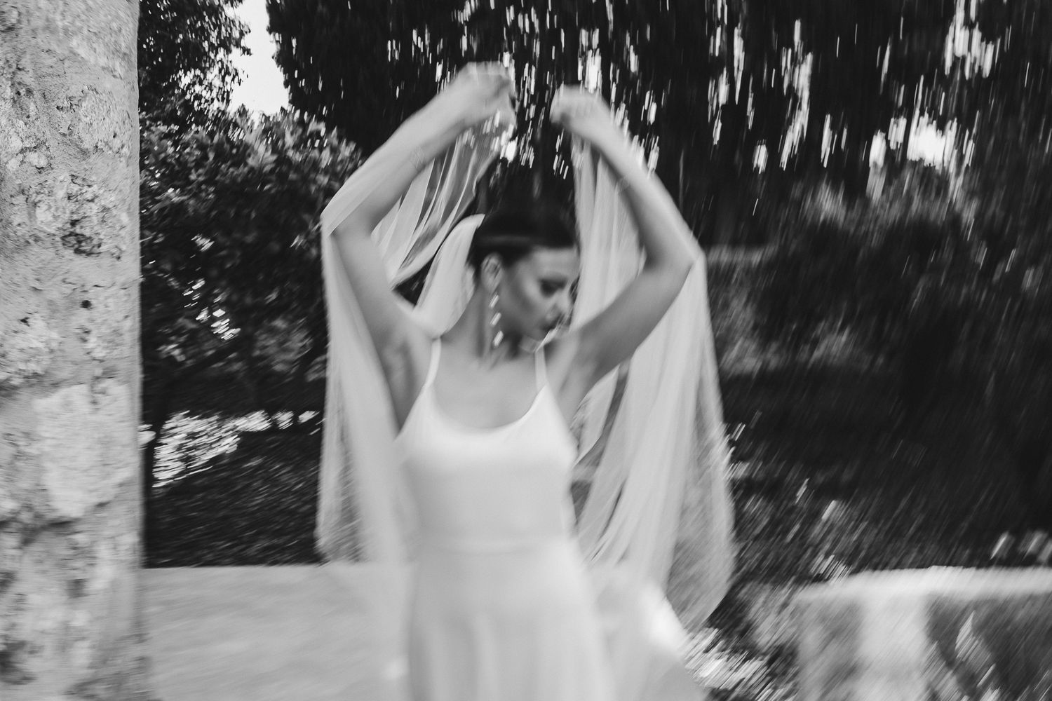 A bride in a white dress adjusts her flowing veil outdoors in a black and white photograph.