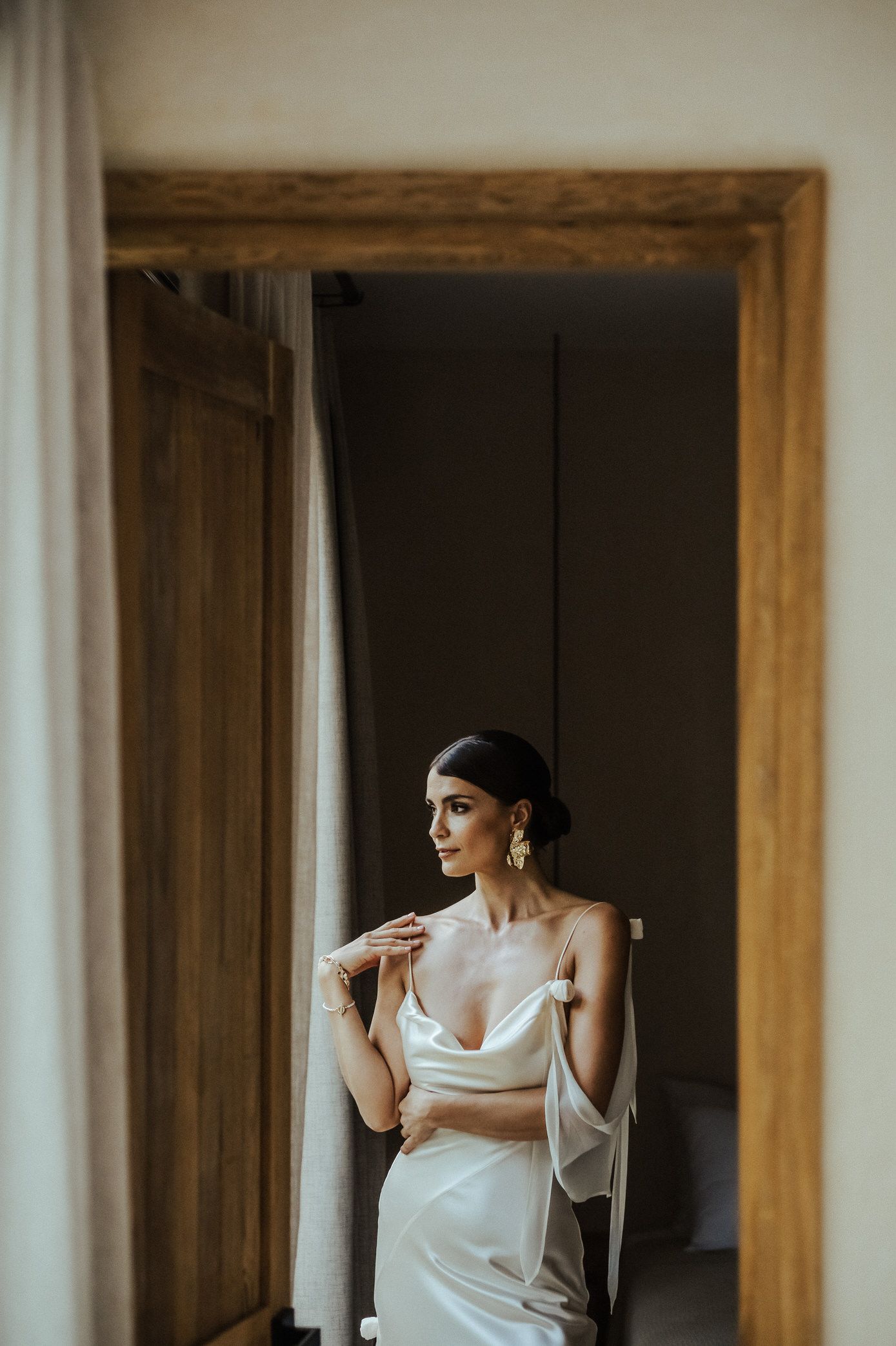 Elegant bride in white silk gown stands thoughtfully by wooden doorframe in soft natural light.