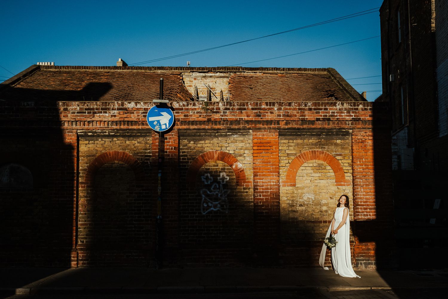 Bride in white dress poses in dramatic light against rustic brick wall during outdoor wedding photoshoot.
