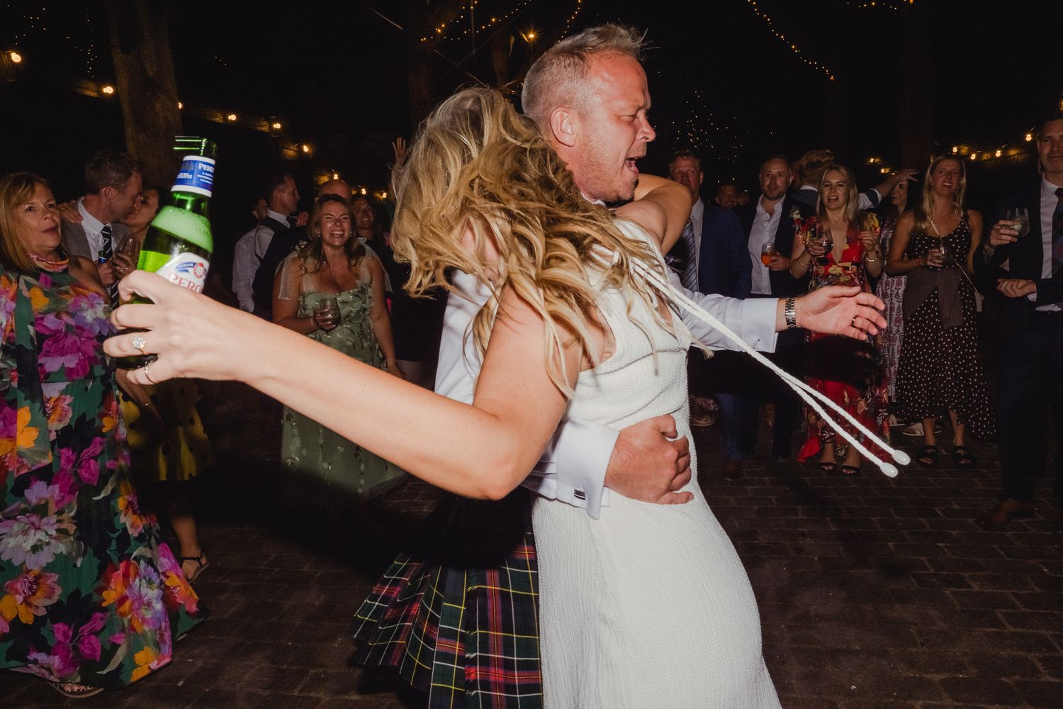 Wedding guests dancing and celebrating on a dark dance floor illuminated by evening lights.