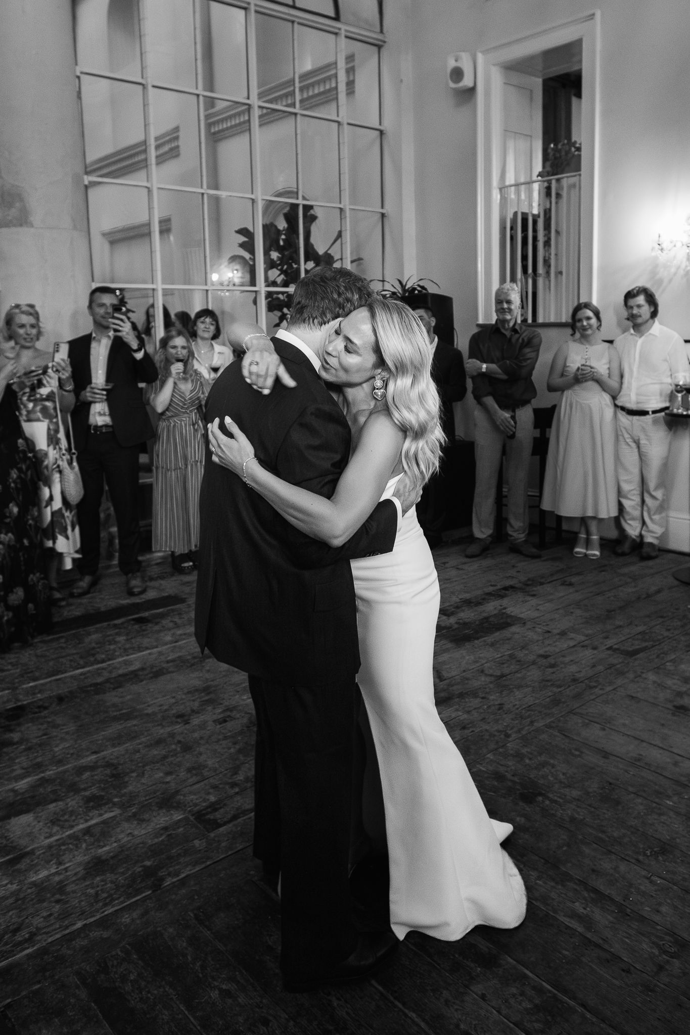 Black and white photo of a bride in a fitted gown embracing her groom during a first dance as wedding guests watch in a historic ballroom.