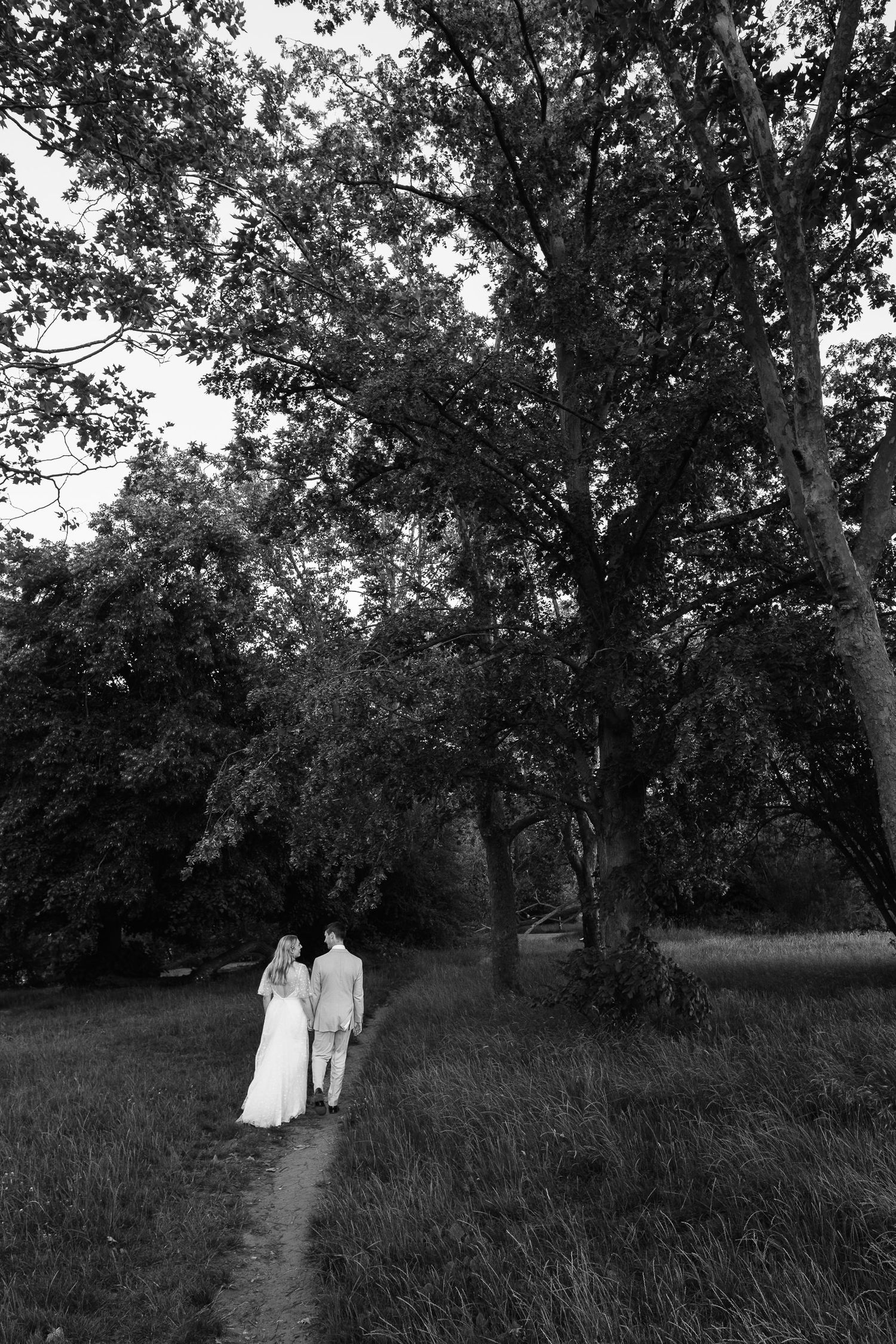 Bride in a long white gown and groom in a light suit walk hand in hand along a grassy path beneath tall trees in black and white.