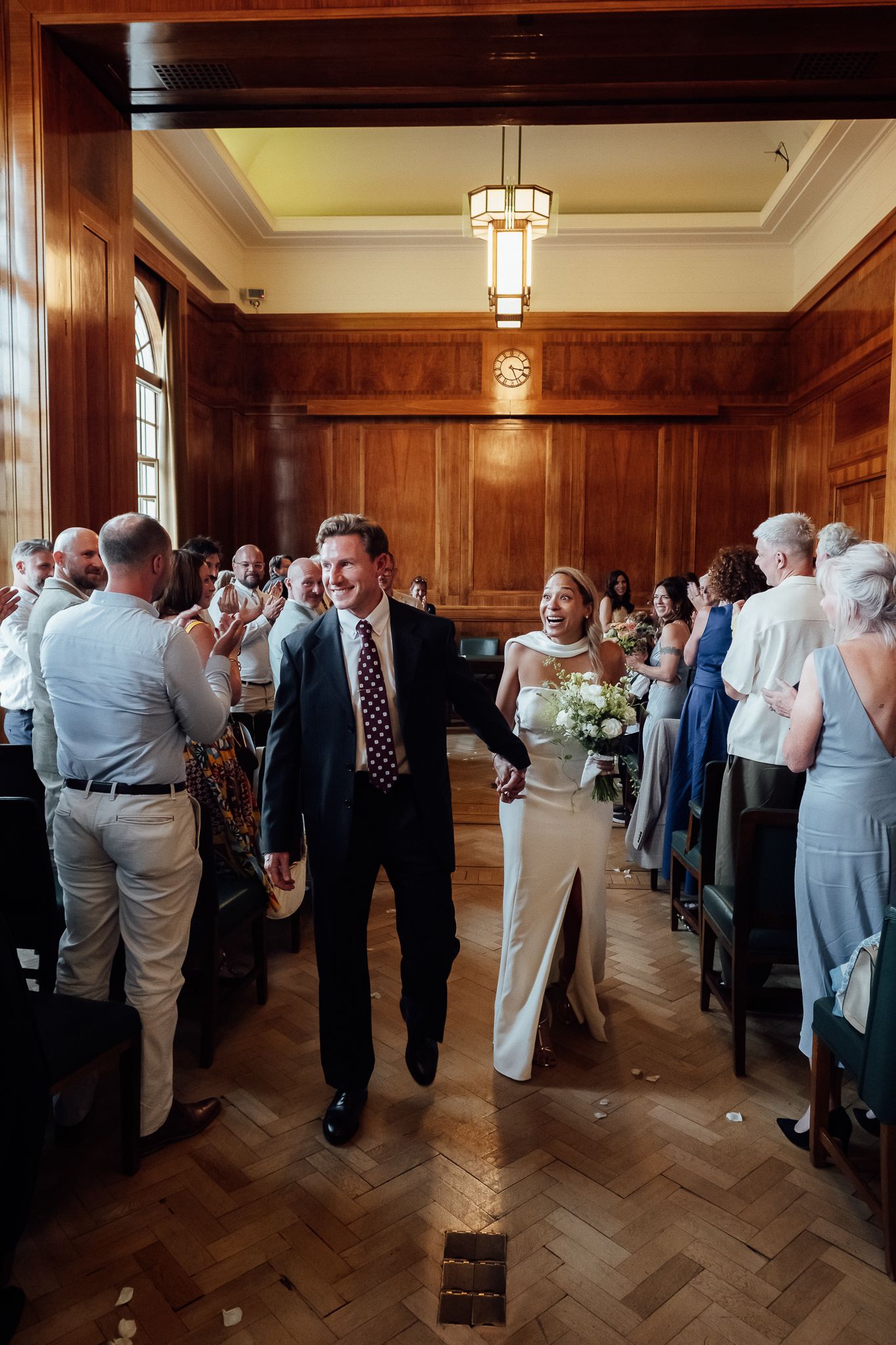 Bride and groom walk down the aisle hand in hand as guests applaud in a wood-paneled wedding ceremony room with a herringbone floor.