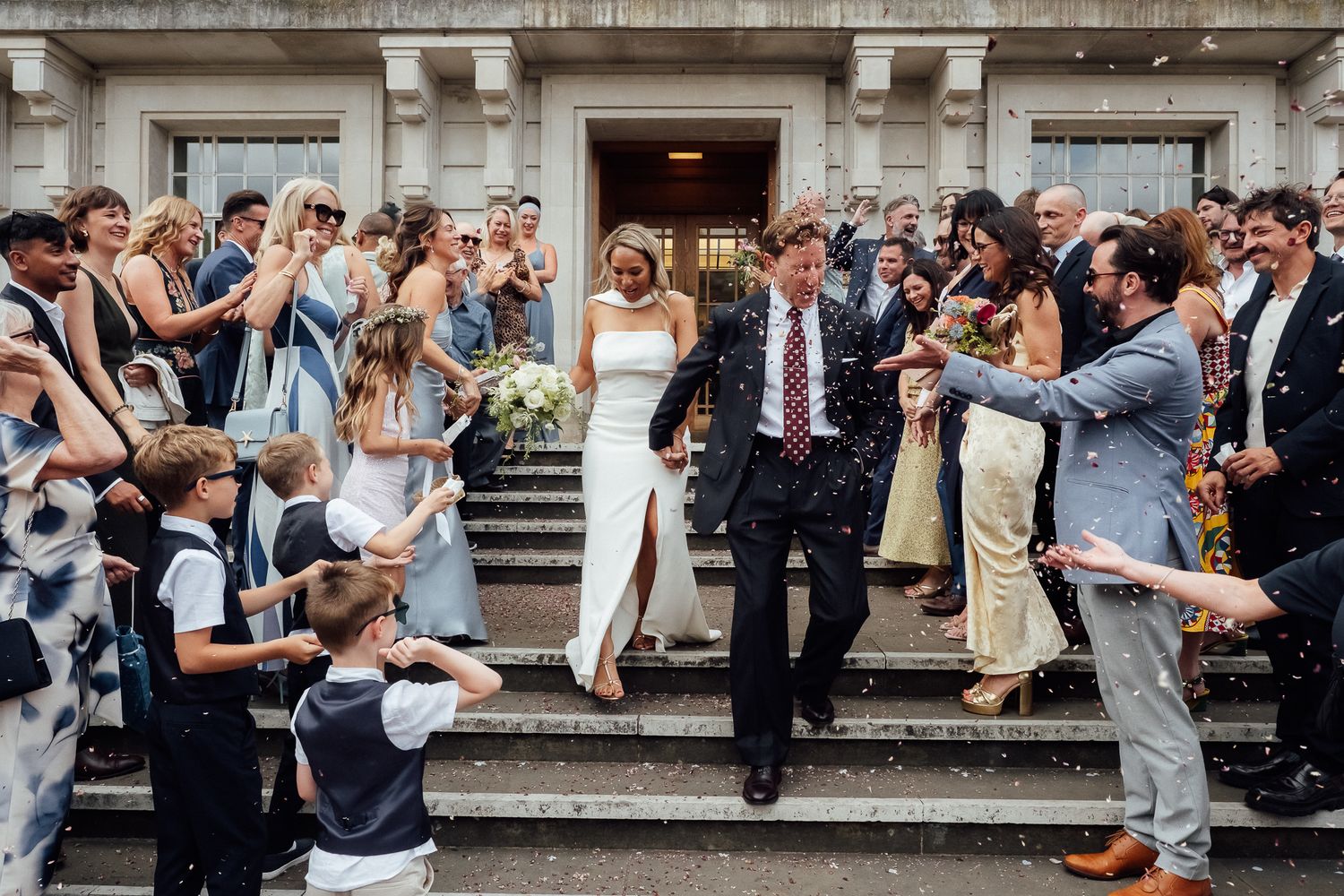 Bride and groom walk down stone steps outside a grand building as smiling wedding guests and children cheer and celebrate around them.