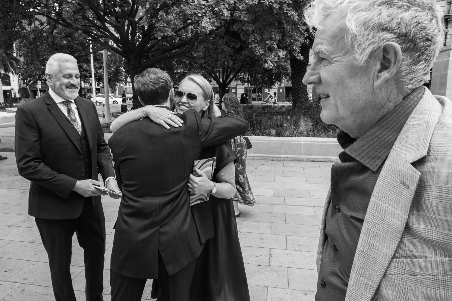 Black and white photo of a man in a suit embracing a woman in sunglasses outdoors while two other men look on.