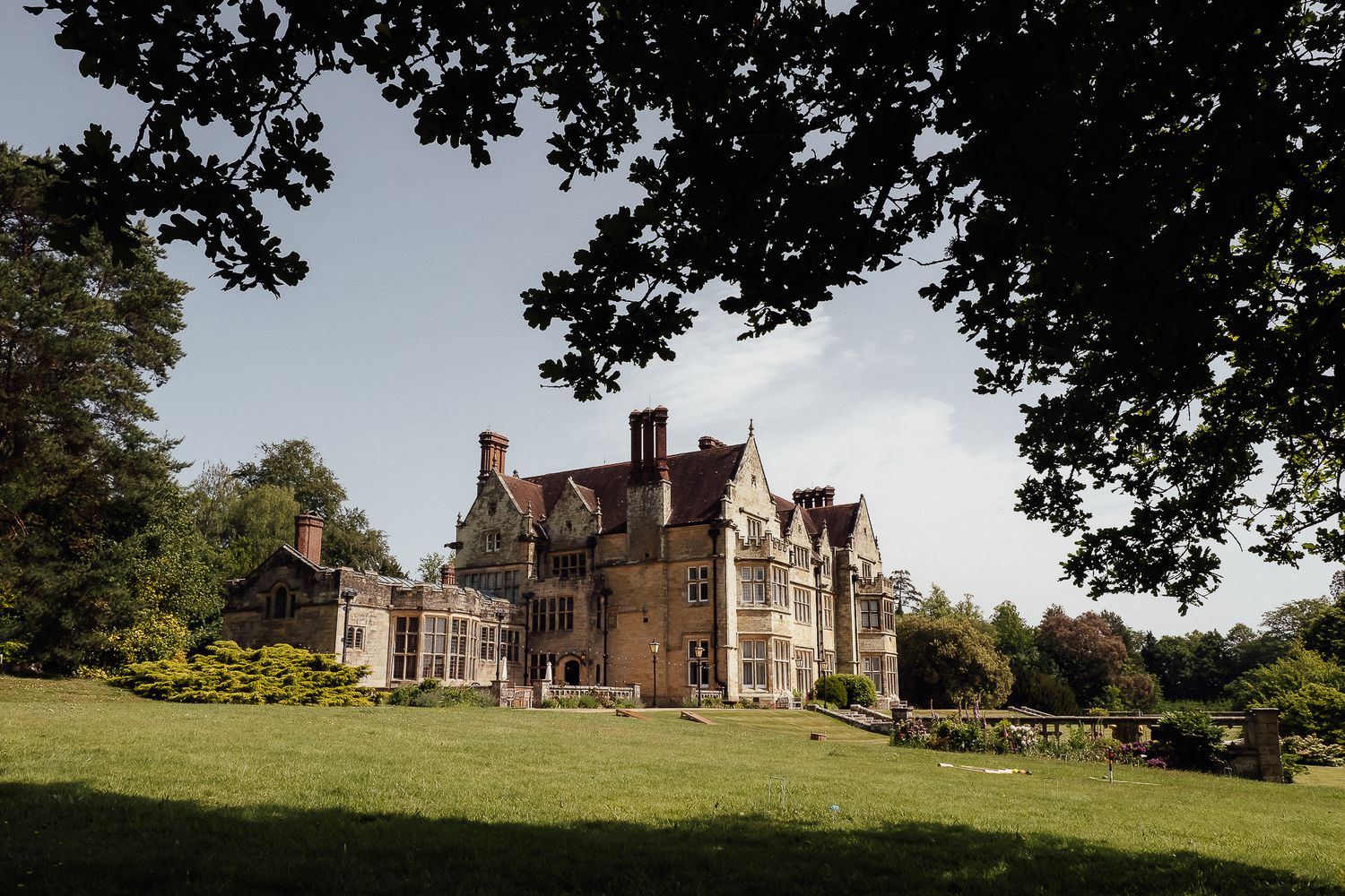 Grand Victorian manor house surrounded by lush green lawns and mature oak trees under a bright blue sky.