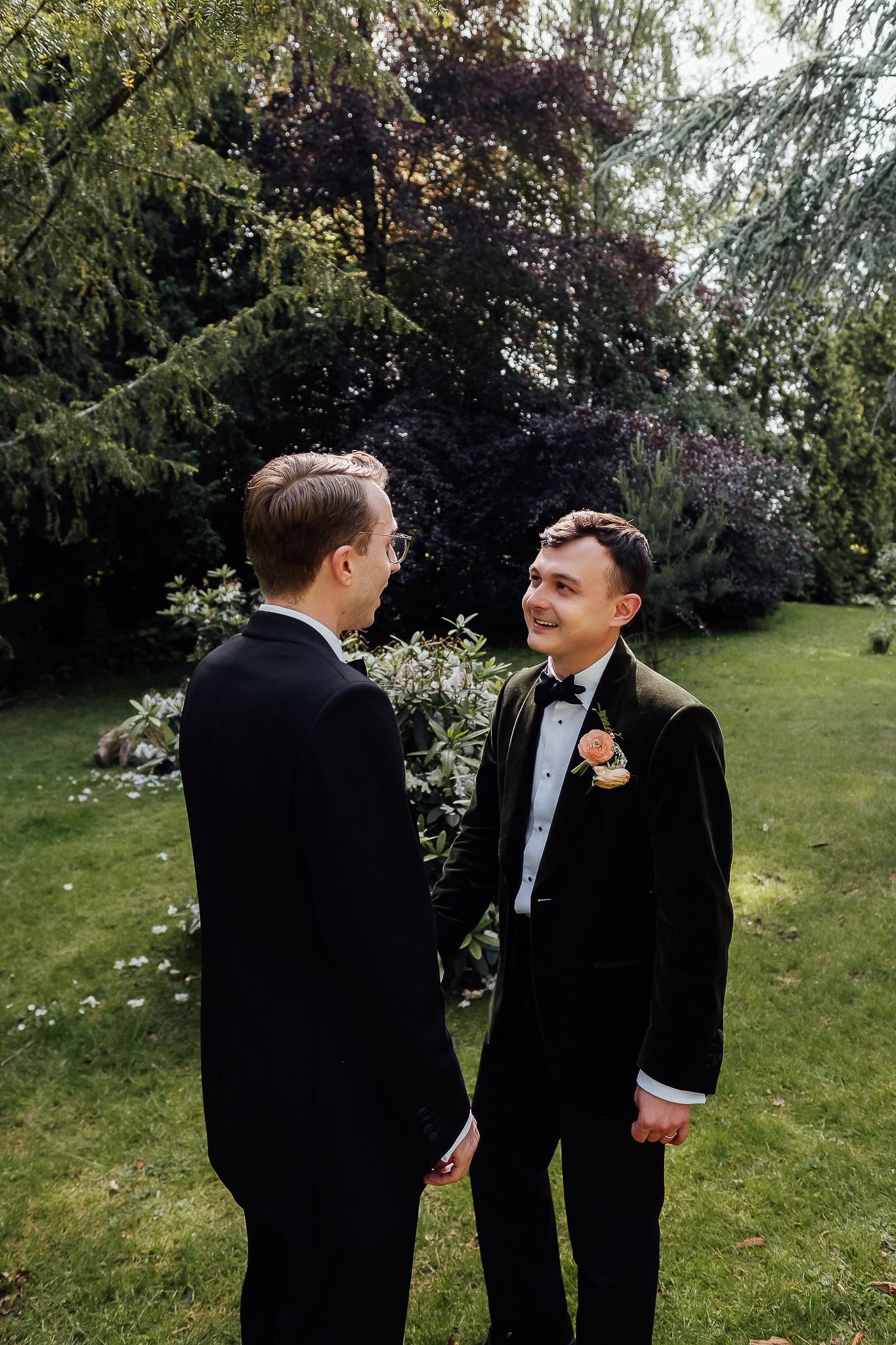 Two grooms in black tuxedos share a moment outdoors in a lush garden setting on their wedding day.