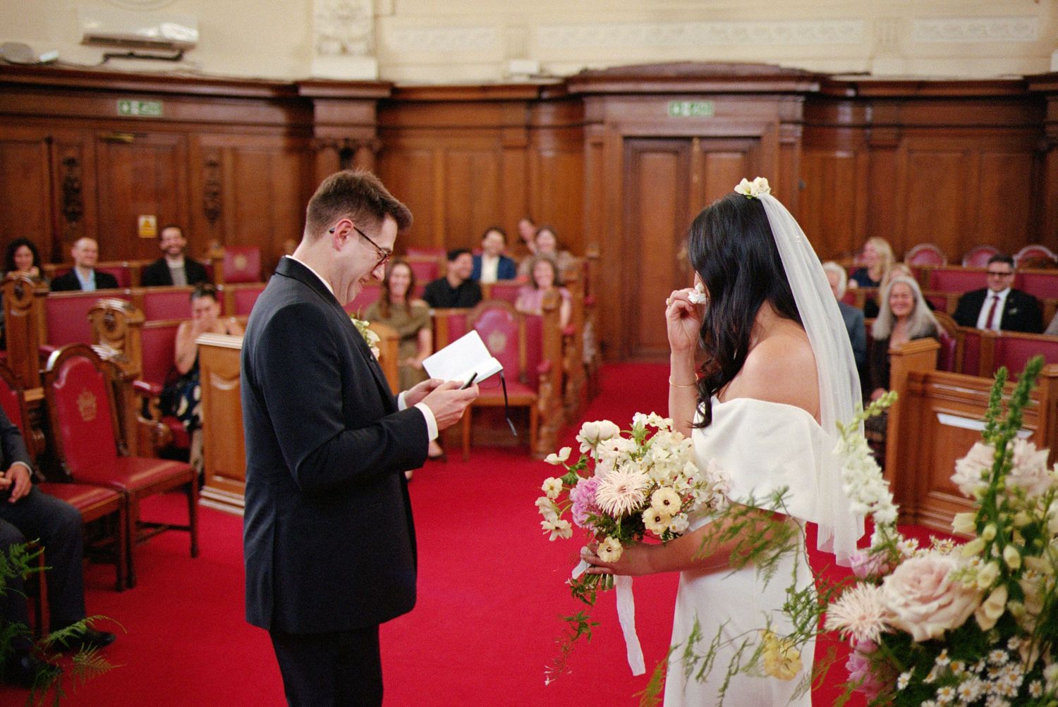 Groom reads vows to bride holding floral bouquet during intimate civil wedding ceremony in elegant wood-paneled hall.