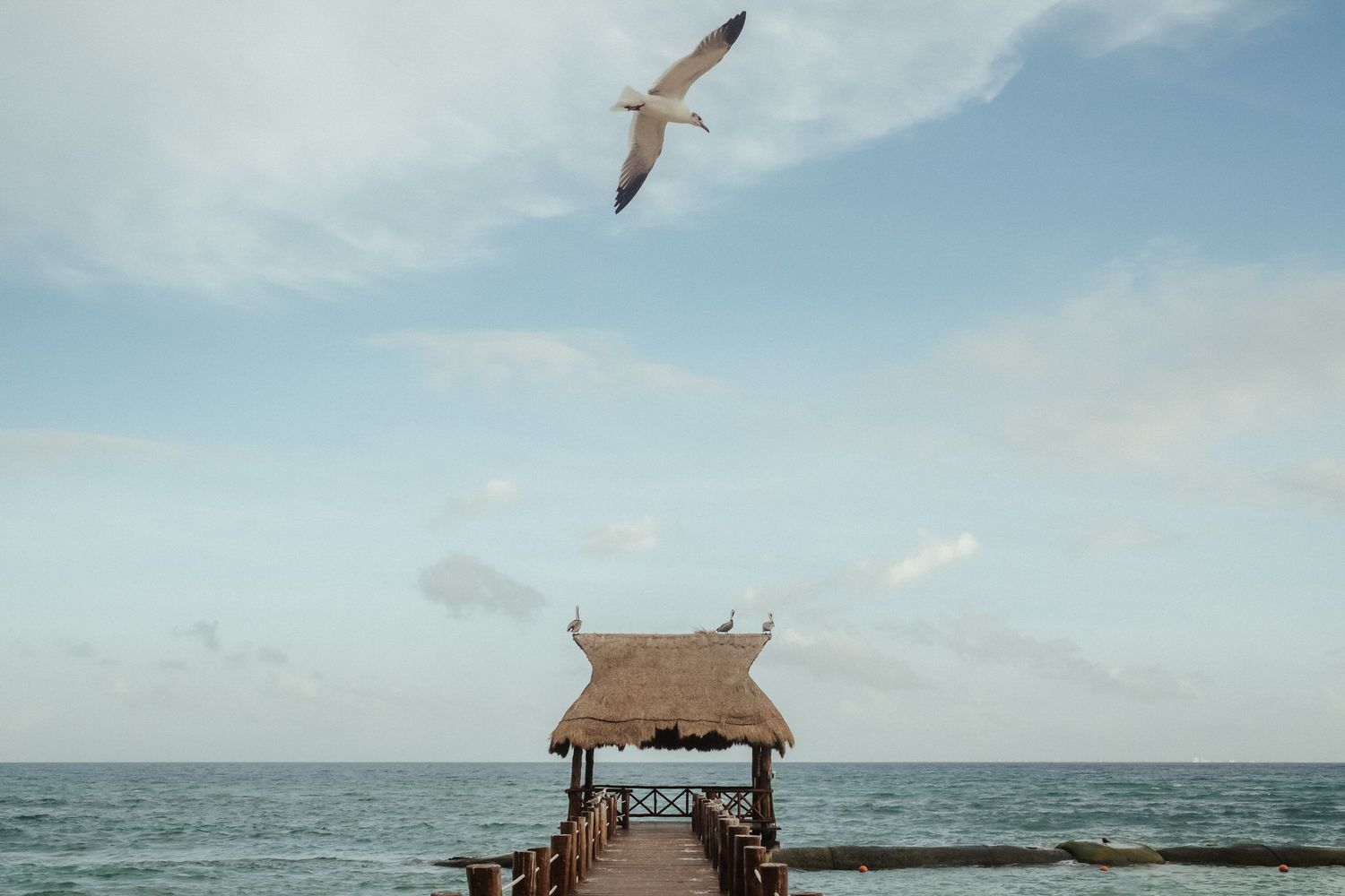 A scenic beachfront tiki hut stands on wooden stilts overlooking the turquoise ocean waters under a cloudy sky.