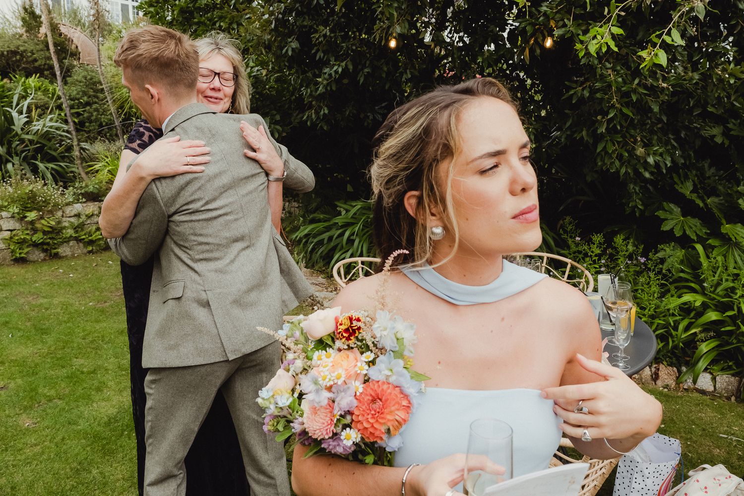 Wedding guests share emotional embraces in a garden setting with a bouquet of peach and coral flowers.
