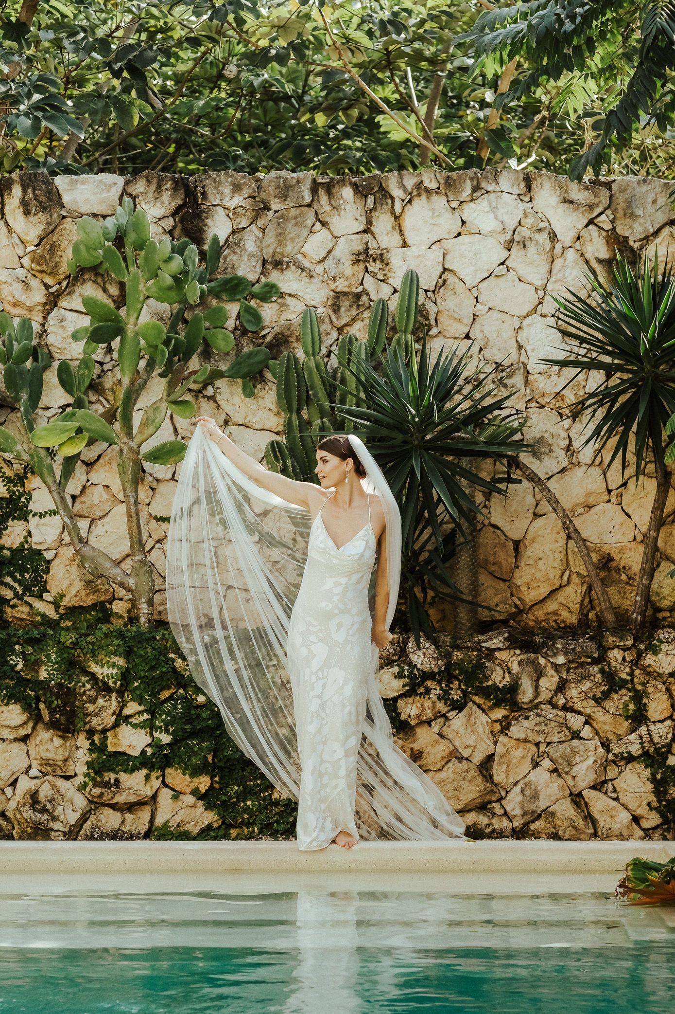 Elegant bride in white lace gown poses gracefully by a luxurious stone-walled pool surrounded by tropical plants.