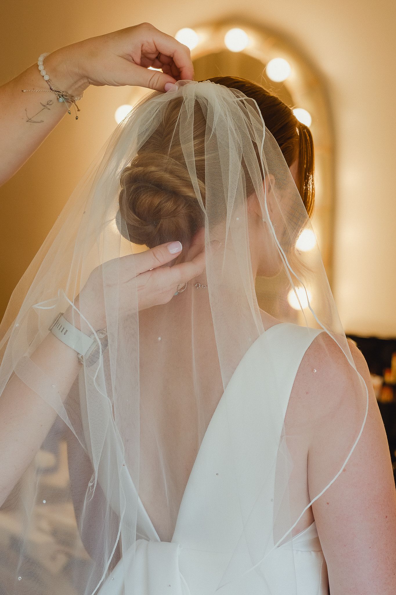 Hands adjusting a delicate white wedding veil on a bride wearing a sleeveless dress in warm lighting.