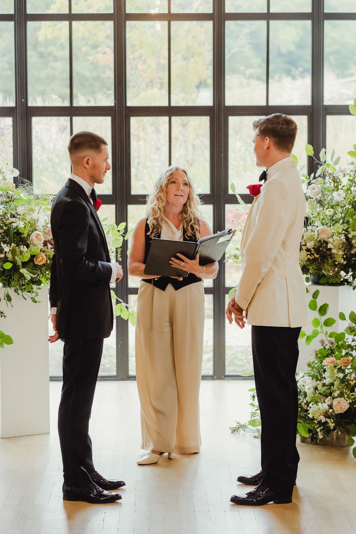 Wedding party members in formal attire line up by large windows in a bright venue with floral arrangements.