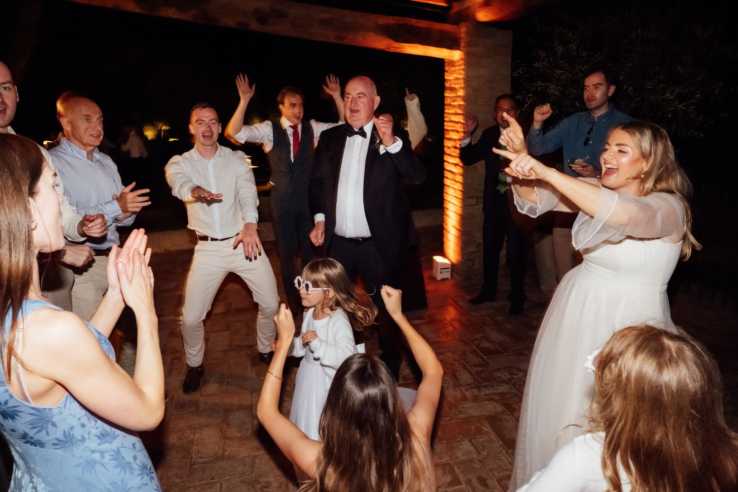 Wedding guests dance and clap joyfully at a nighttime reception as the bride and children celebrate on the dance floor.