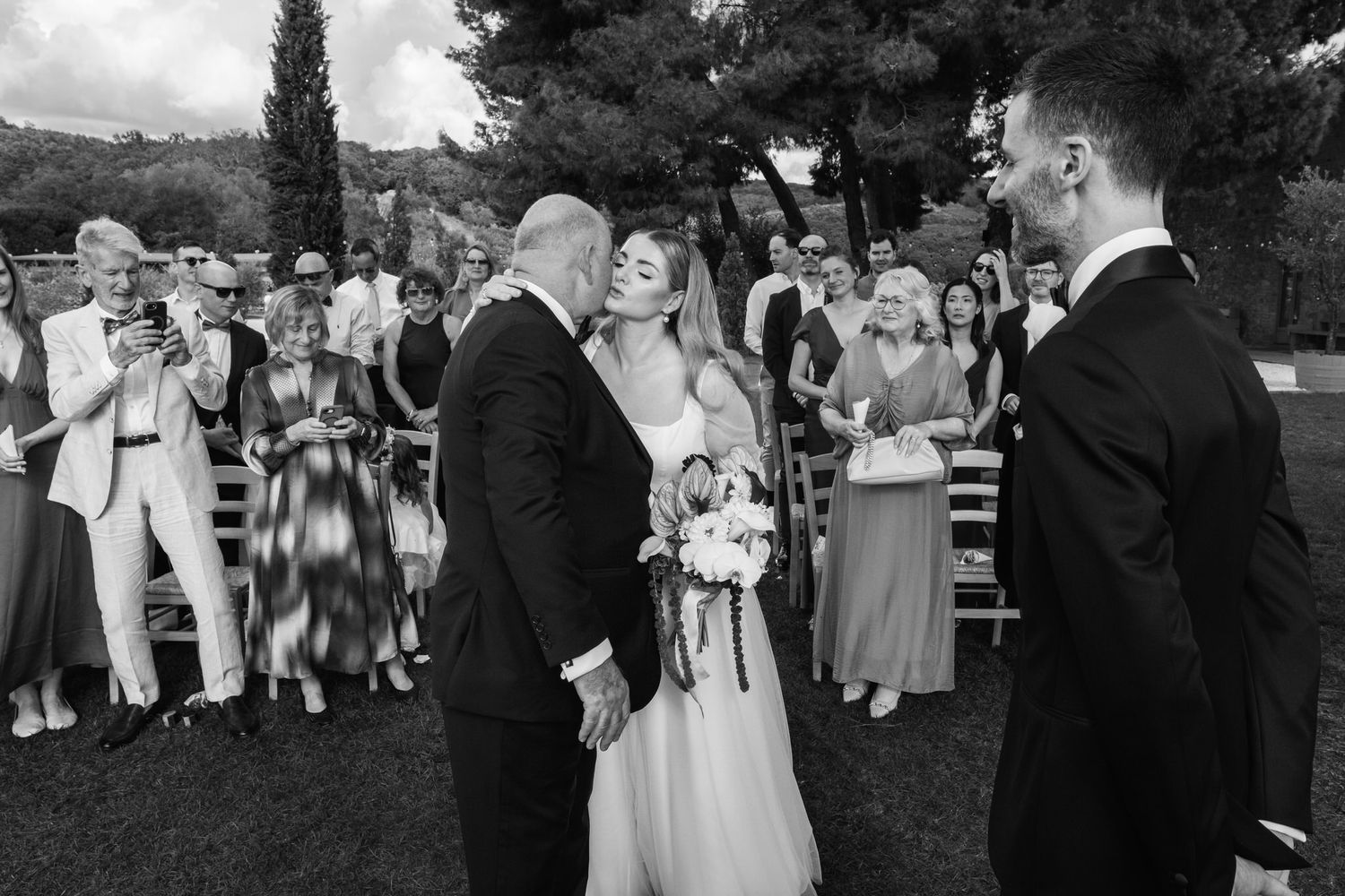 Black and white wedding photo of a bride in a lace gown with bouquet kissing an older man on the cheek as guests watch outdoors.
