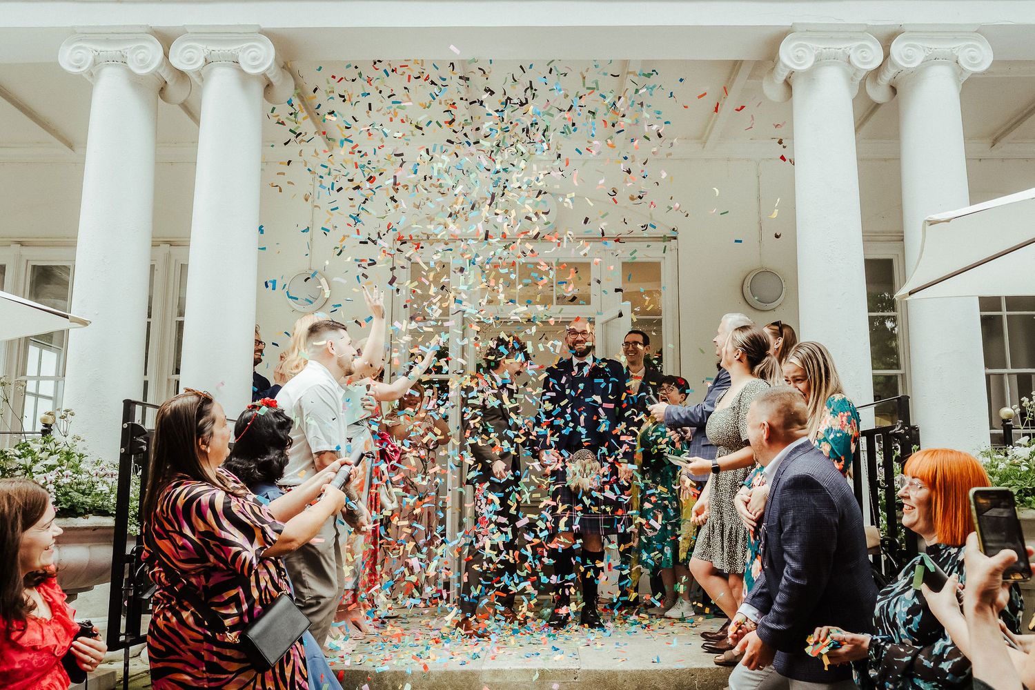 Wedding guests throw colorful confetti over a newlywed couple walking out beneath white columns at a sunlit celebration.