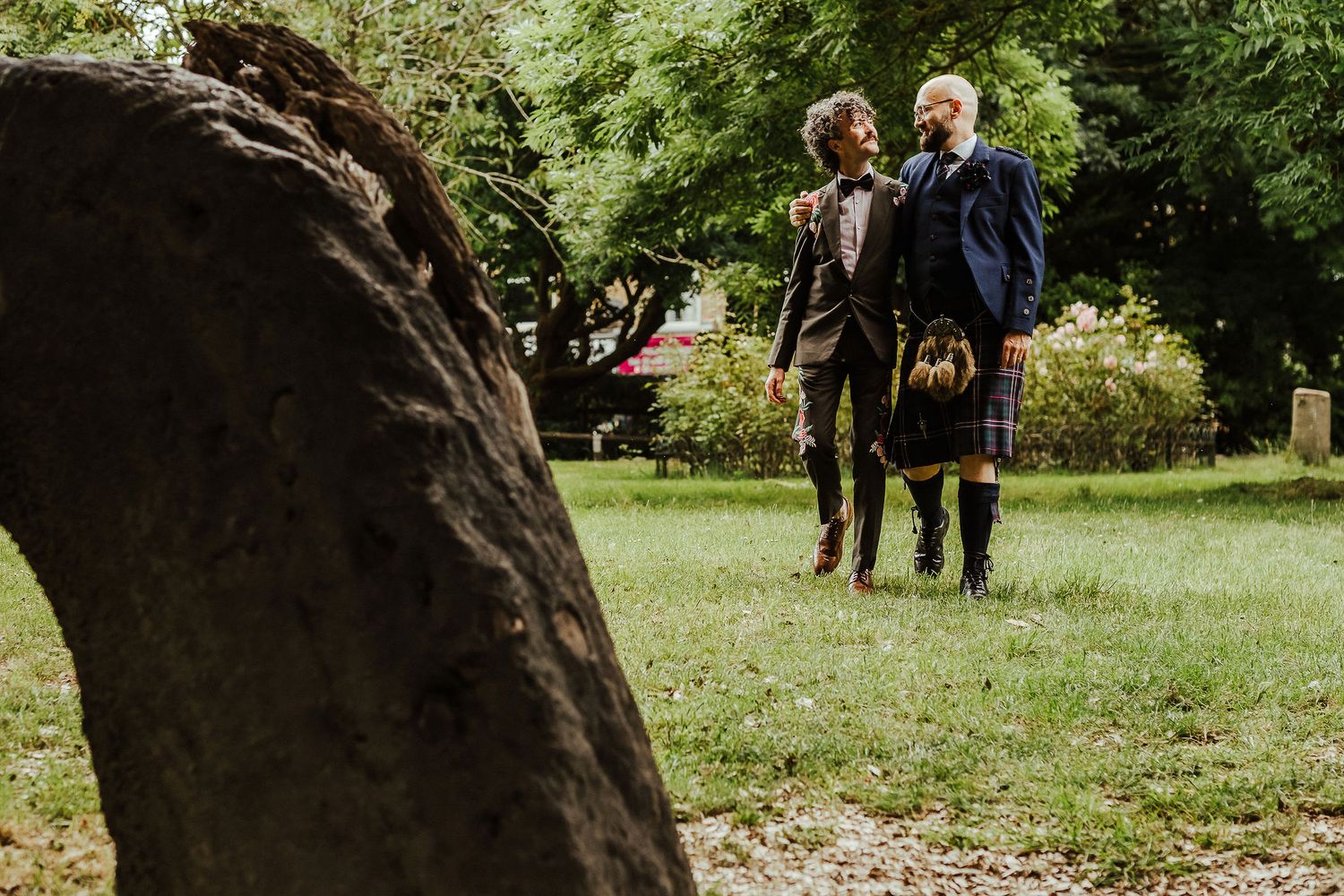 Two people in formal Scottish attire, one wearing a kilt, stand together on a sunny grassy lawn beside a large tree trunk in the foreground.