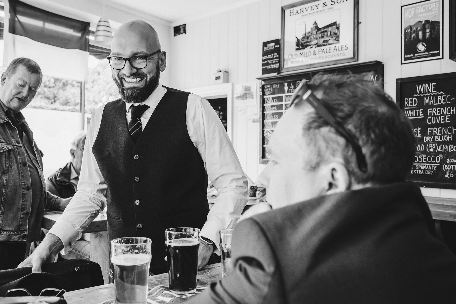 Smiling bald bearded man in a vest and tie chats with seated patrons over pints of beer and stout in a cozy pub.