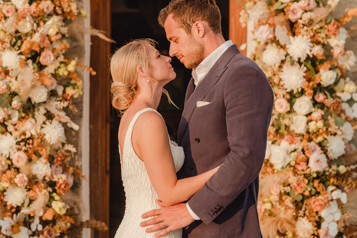 A couple shares an intimate moment during their wedding in front of a backdrop of peach and cream colored floral arrangements.