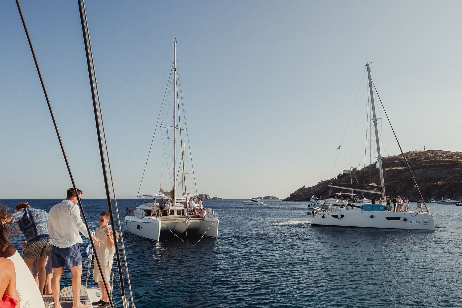 Sailboats anchored in a calm blue bay with coastal landscape and clear skies in the background.