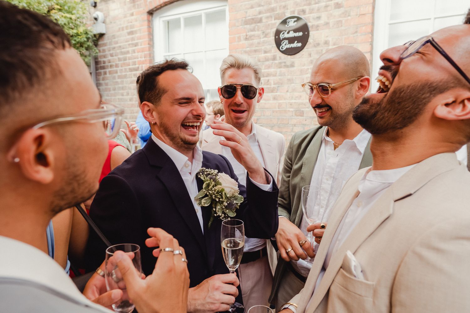 A group of wedding guests laugh and socialize during an outdoor reception near a brick building.