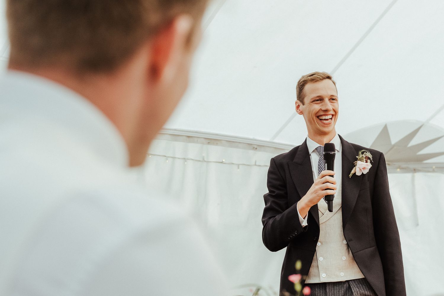 Wedding reception attendees share laughter during heartfelt speeches under a white tent.