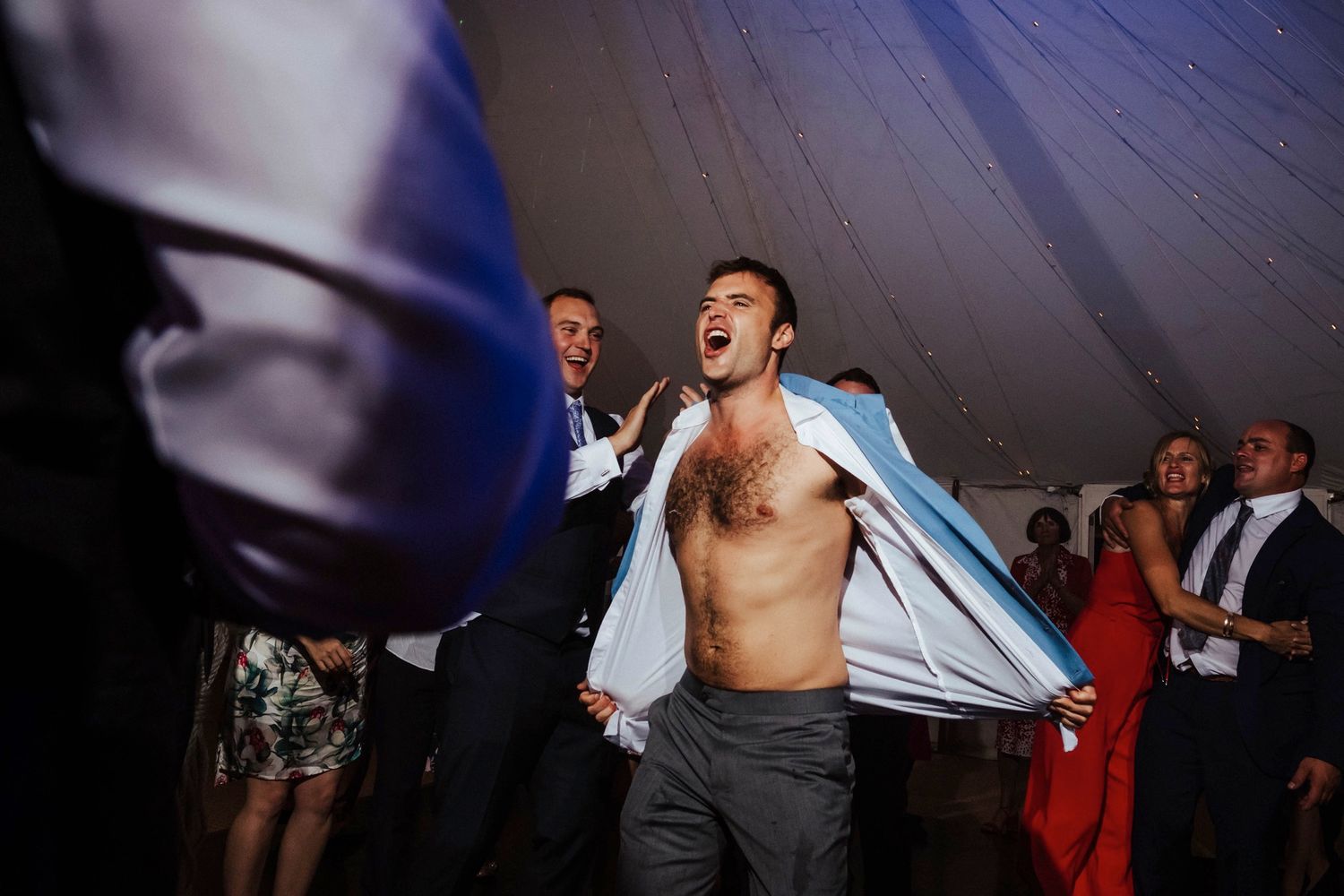 Shirtless guest dances enthusiastically at wedding reception under white tent.