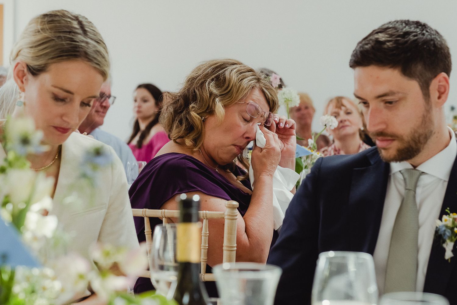 Wedding guests enjoy a lively reception dinner at decorated tables with flowers and glassware.