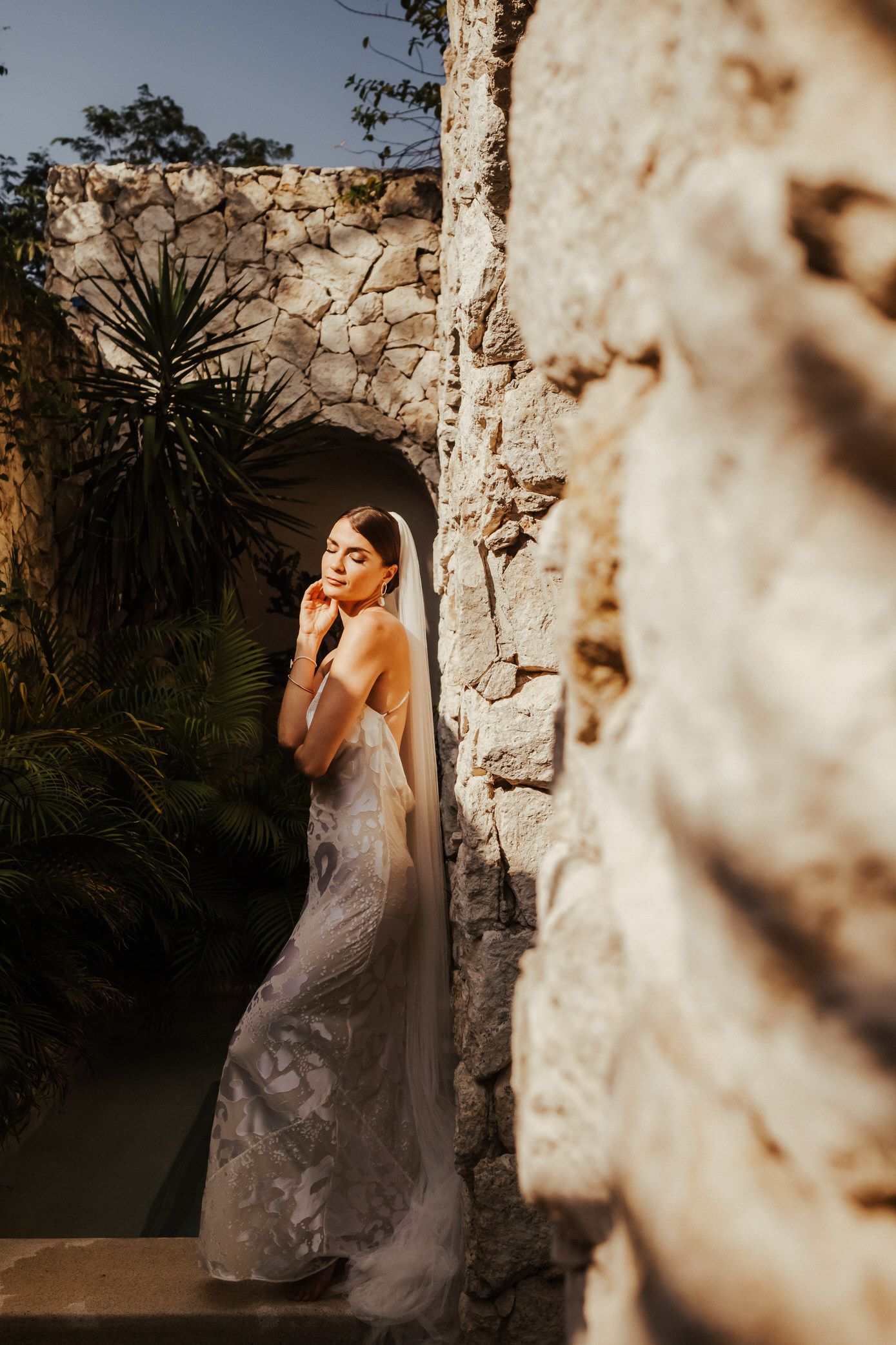 A bride in a silver-gray wedding dress poses gracefully against a rustic stone wall in soft natural lighting.