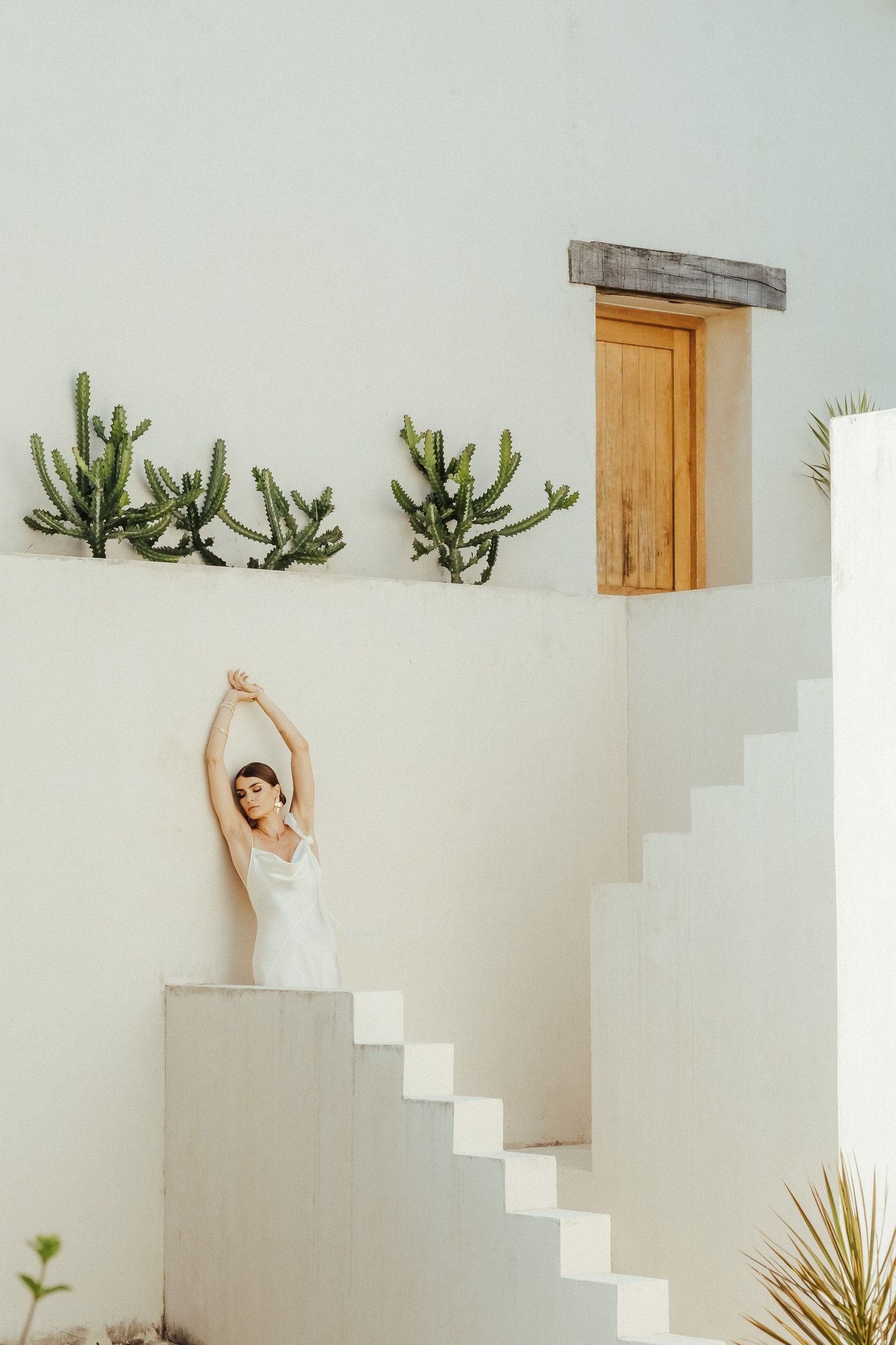 A figure in white stands elegantly against a minimalist white wall with stairs and desert plants.