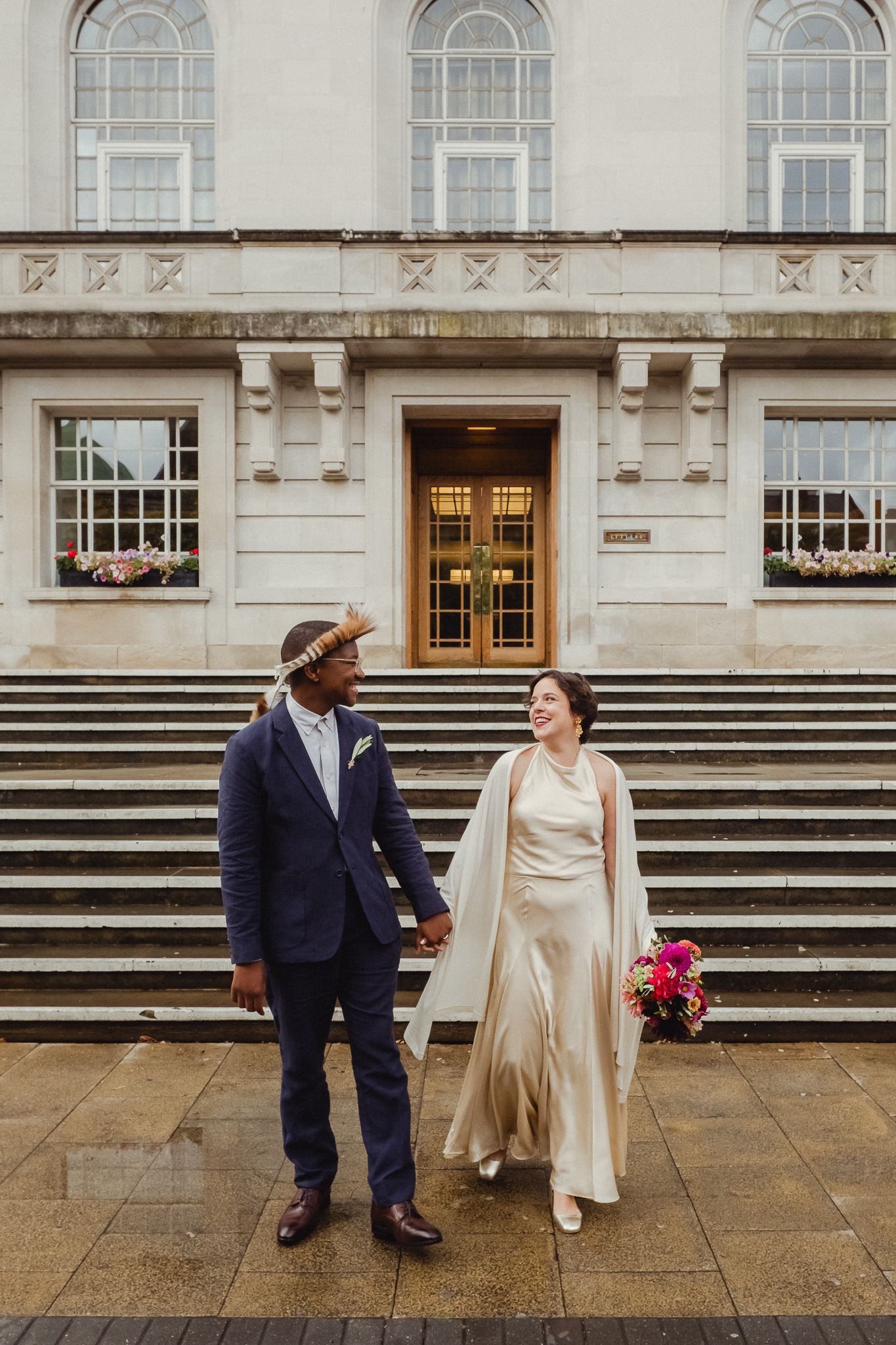 A couple shares a joyful moment on grand stone steps outside an elegant historic building with arched windows.