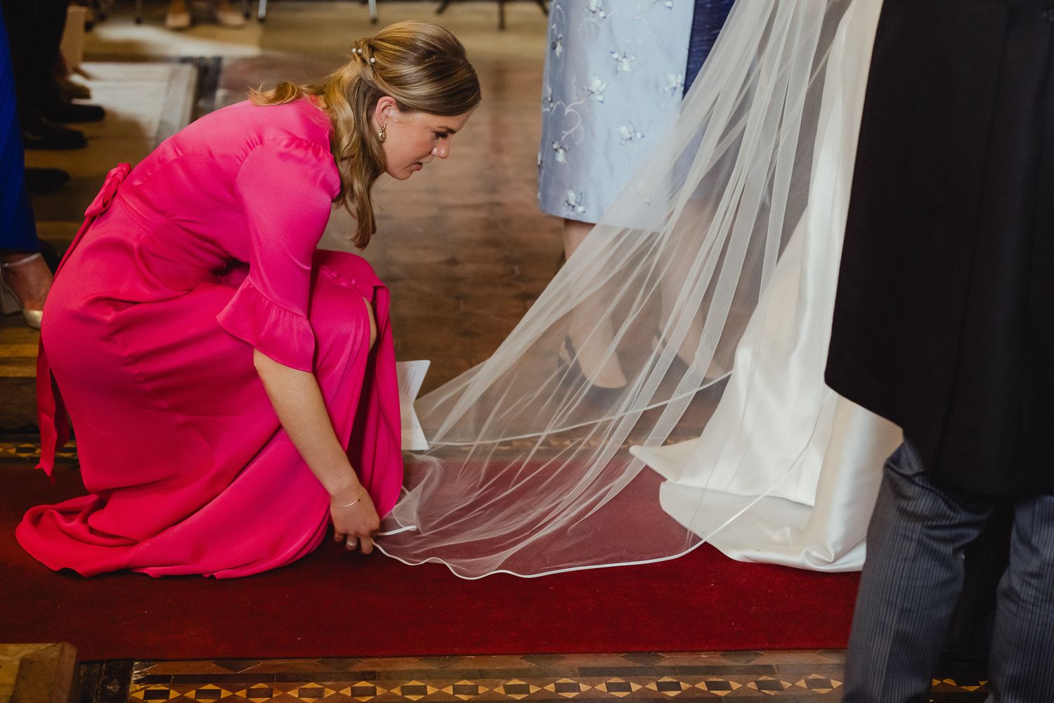 A figure in a bright pink dress helps adjust the hem of a white wedding gown on a wooden floor.