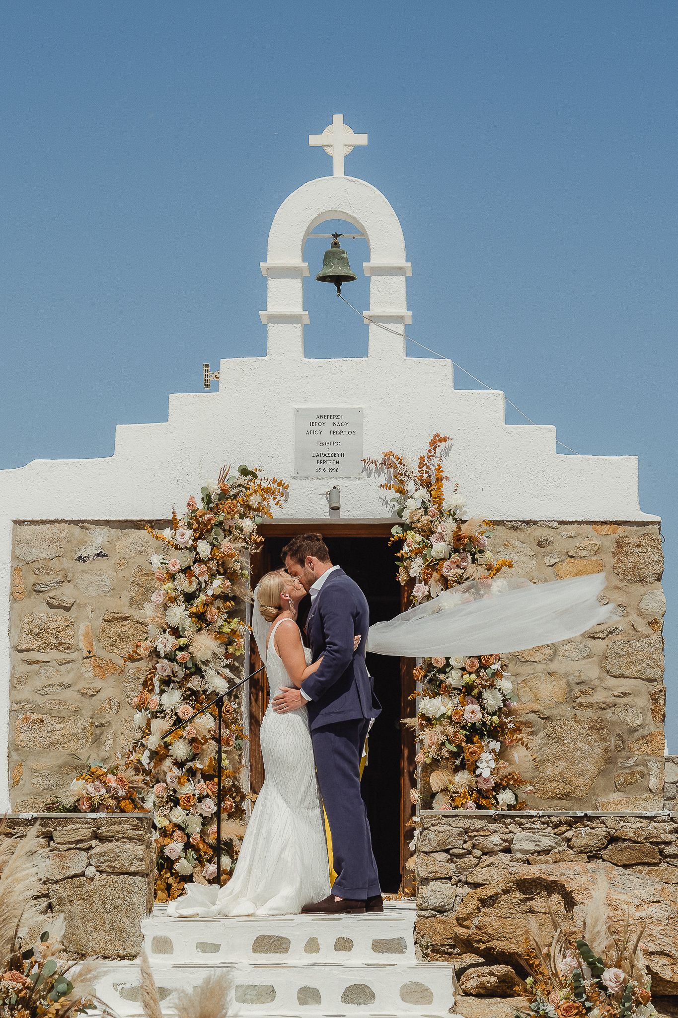 Elegant outdoor wedding ceremony at a whitewashed Mediterranean church with peach and cream floral arch decorations.