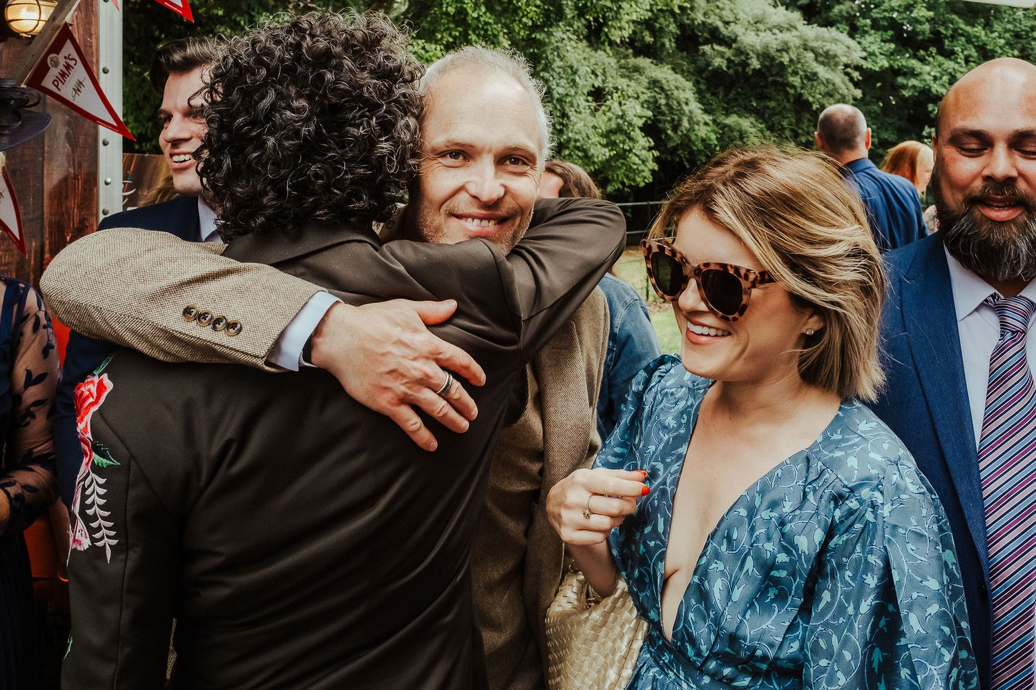 Wedding guests share a warm hug while a smiling woman in sunglasses and a blue floral dress looks on at an outdoor celebration.