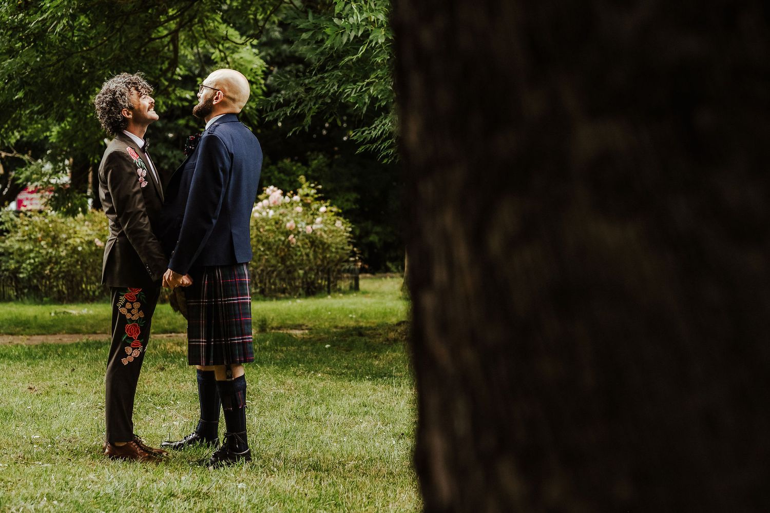 Two grooms in Scottish kilts stand facing each other on a sunlit lawn, framed by trees and garden shrubs, in an outdoor wedding portrait.