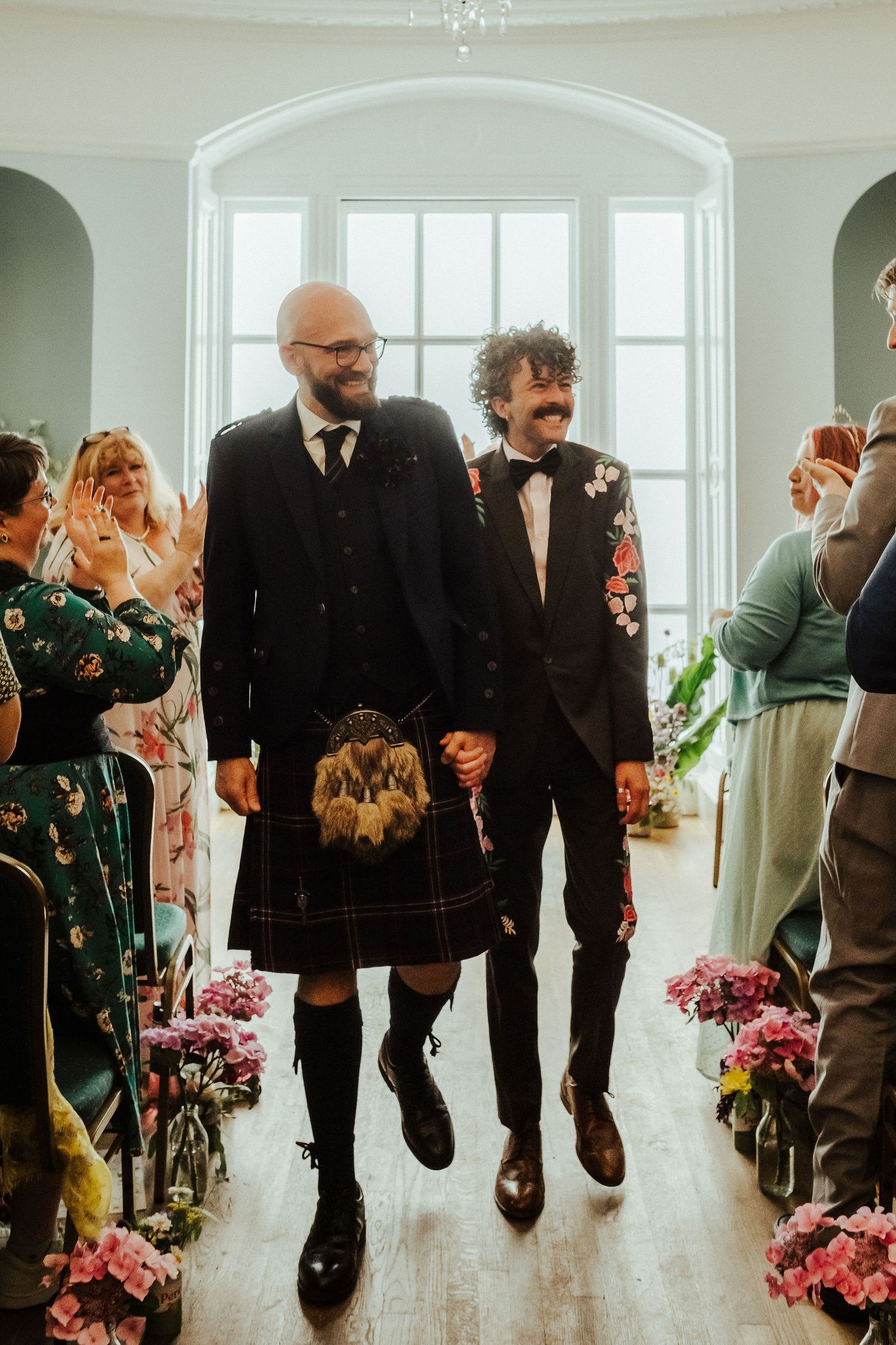 Two grooms holding hands walk down the aisle after their wedding ceremony, one in a kilt, the other in a floral jacket, as guests cheer.