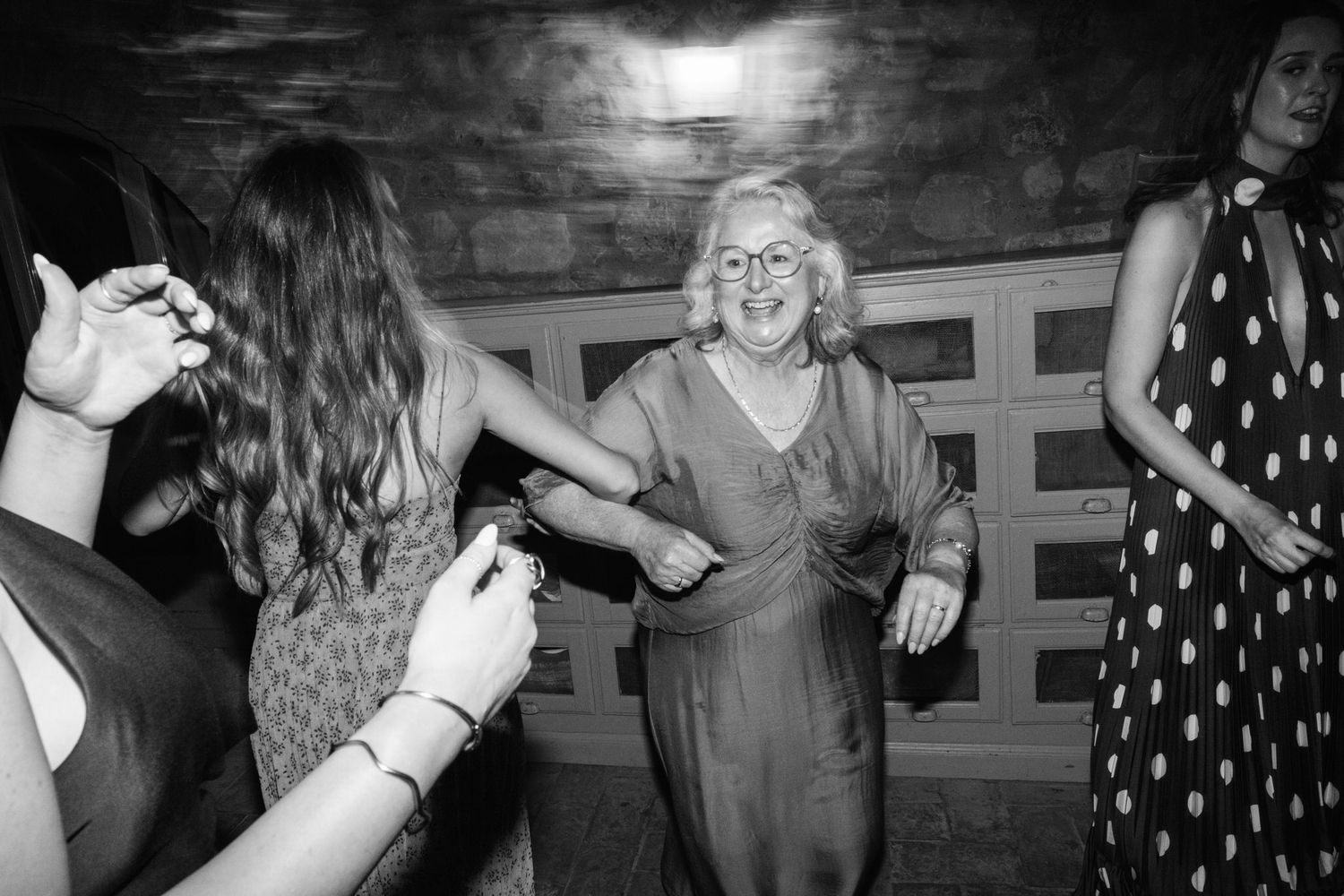 Black and white photo of an older woman with glasses smiling and dancing with guests at a lively indoor party.
