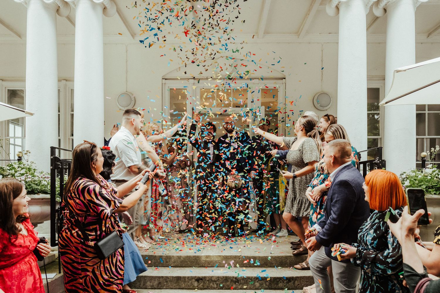 Wedding guests joyfully throw colorful confetti at a couple exiting a bright white venue during their celebration.