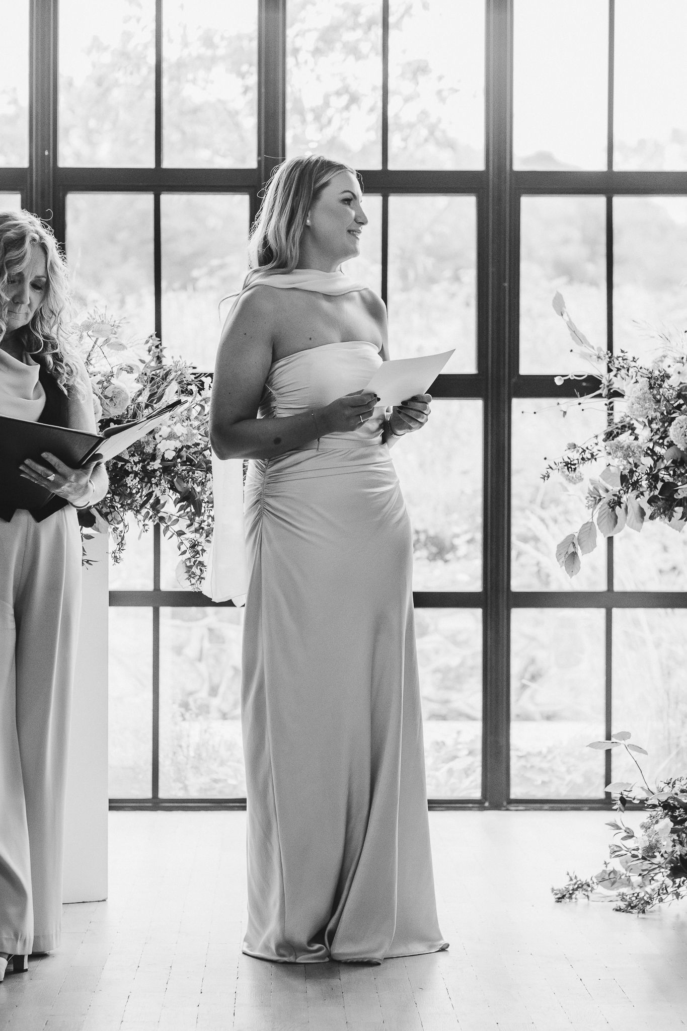 A row of bridesmaids in light-colored dresses hold bouquets while standing by large windows in a bright room.