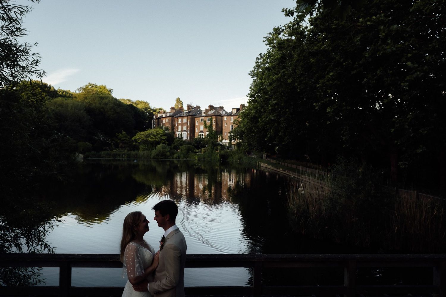 Couple embracing on a wooden bridge at dusk beside a calm lake with a historic manor house and lush green trees in the background.